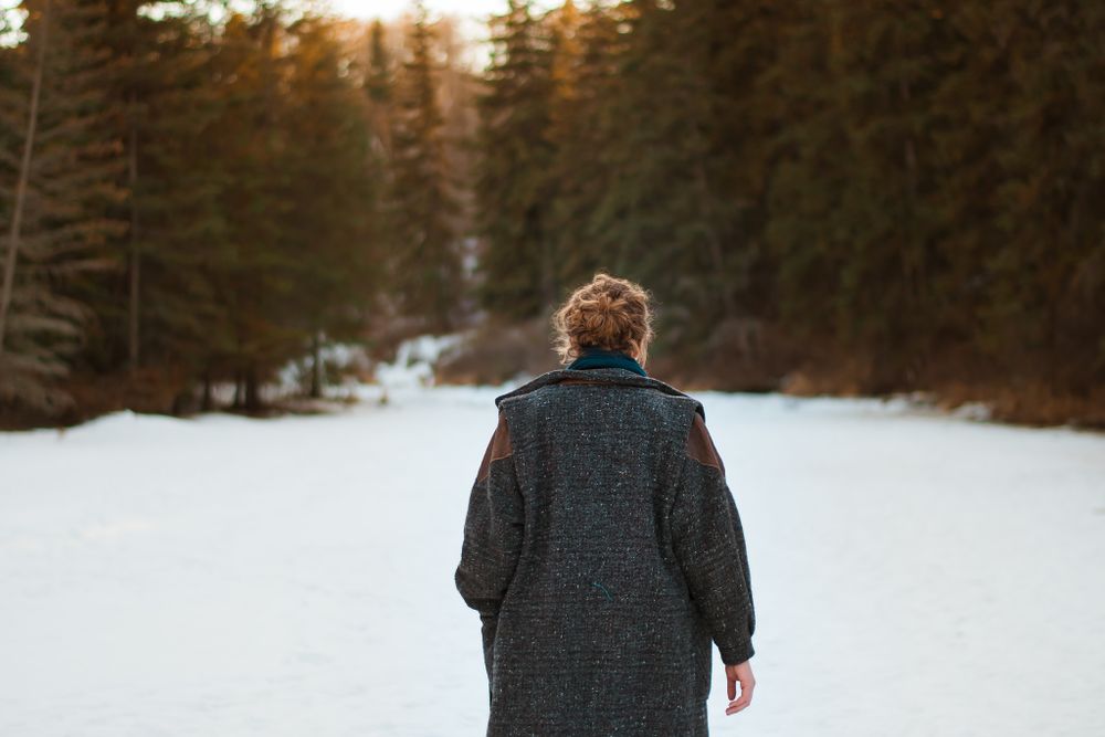 Back view of person in dark wool coat walking in snowy forest landscape.