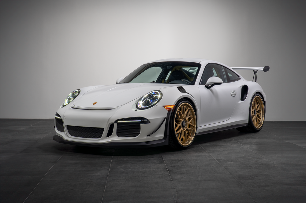 White Porsche 911 GT3 RS sports car with gold wheels sitting on a dark concrete floor against a gray backdrop.