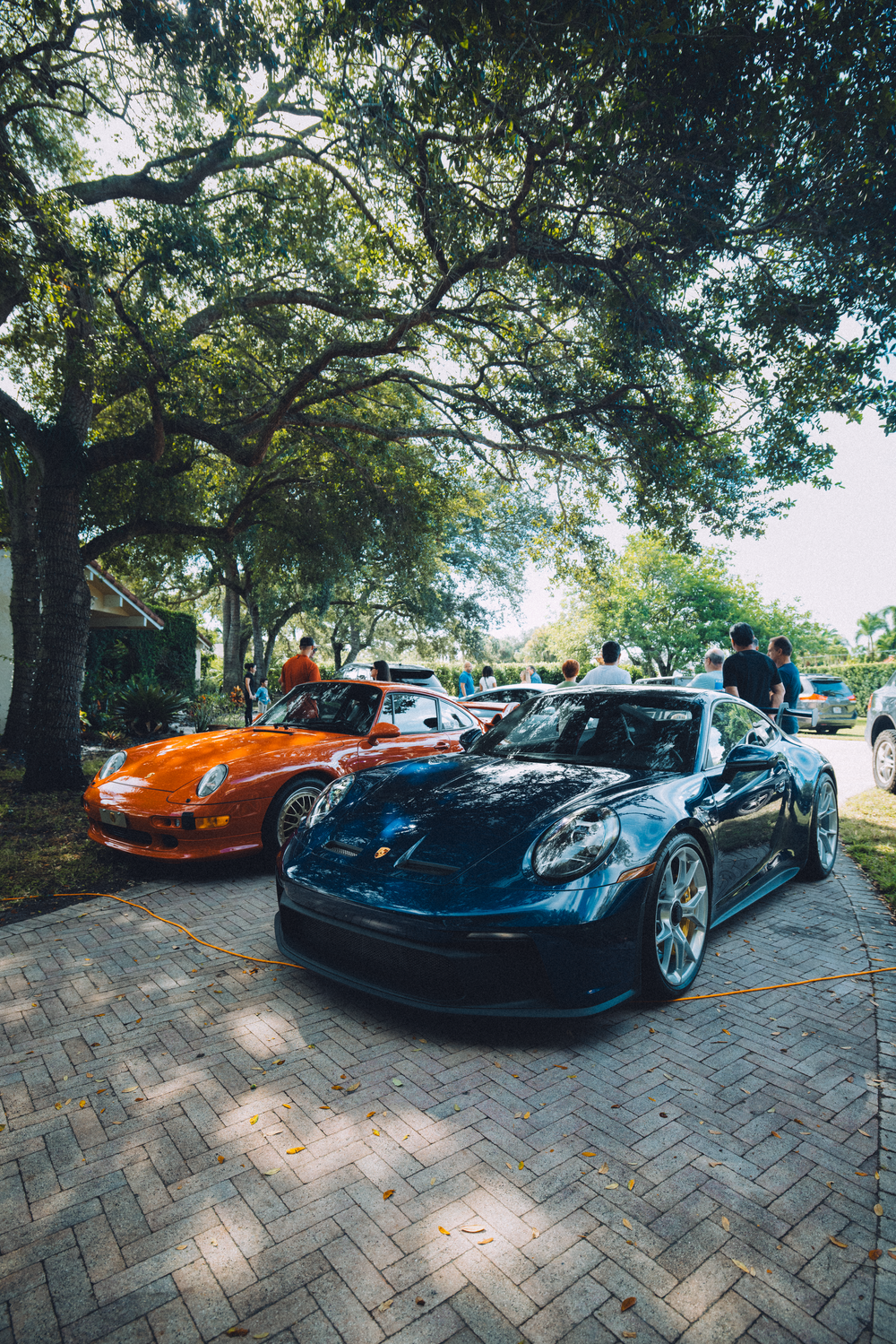 Two luxury Porsche sports cars, a navy GT3 and an orange model, parked on brick pavement under shady trees.