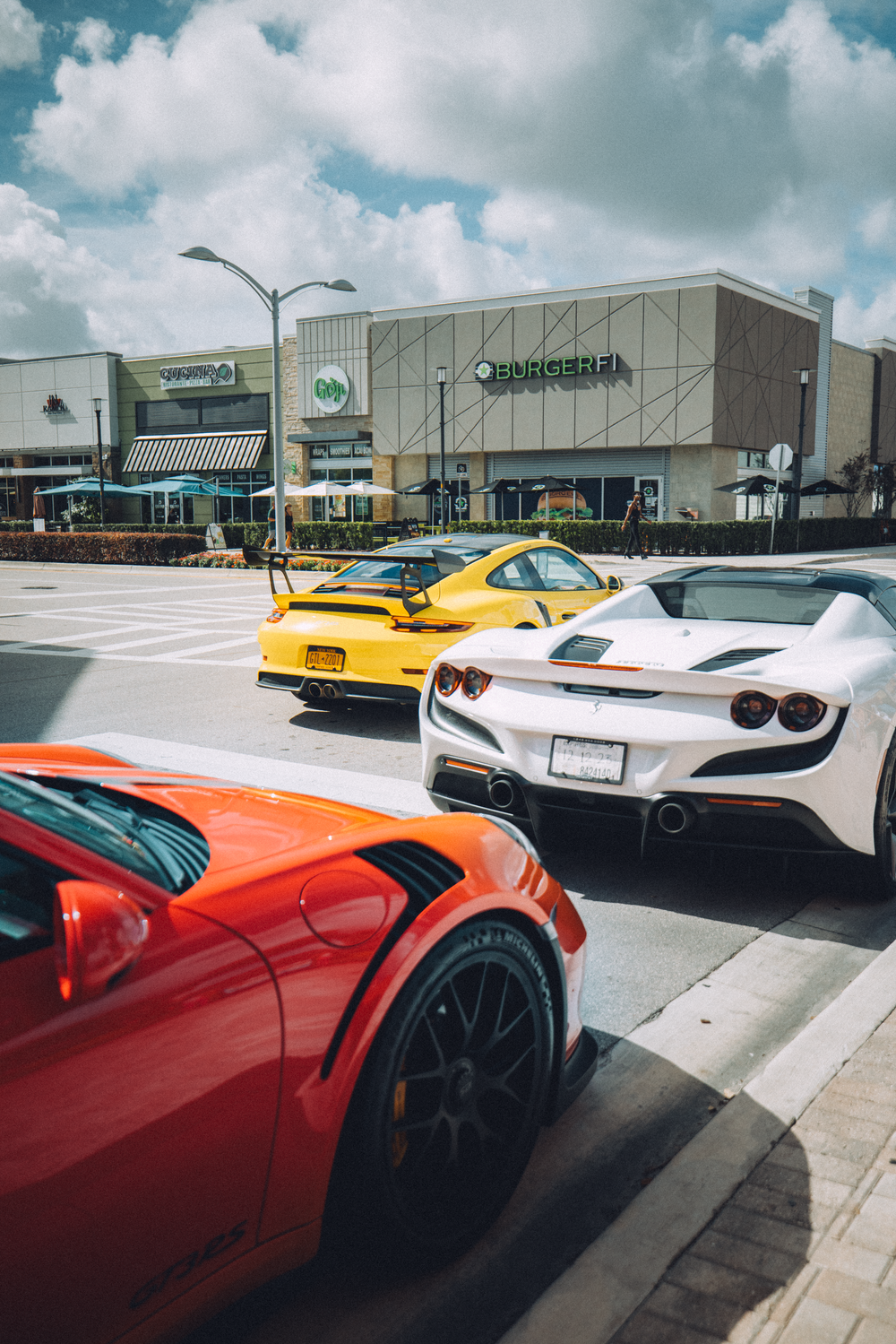 Luxury sports cars including a red, yellow, and white model parked outside a shopping center under cloudy skies.