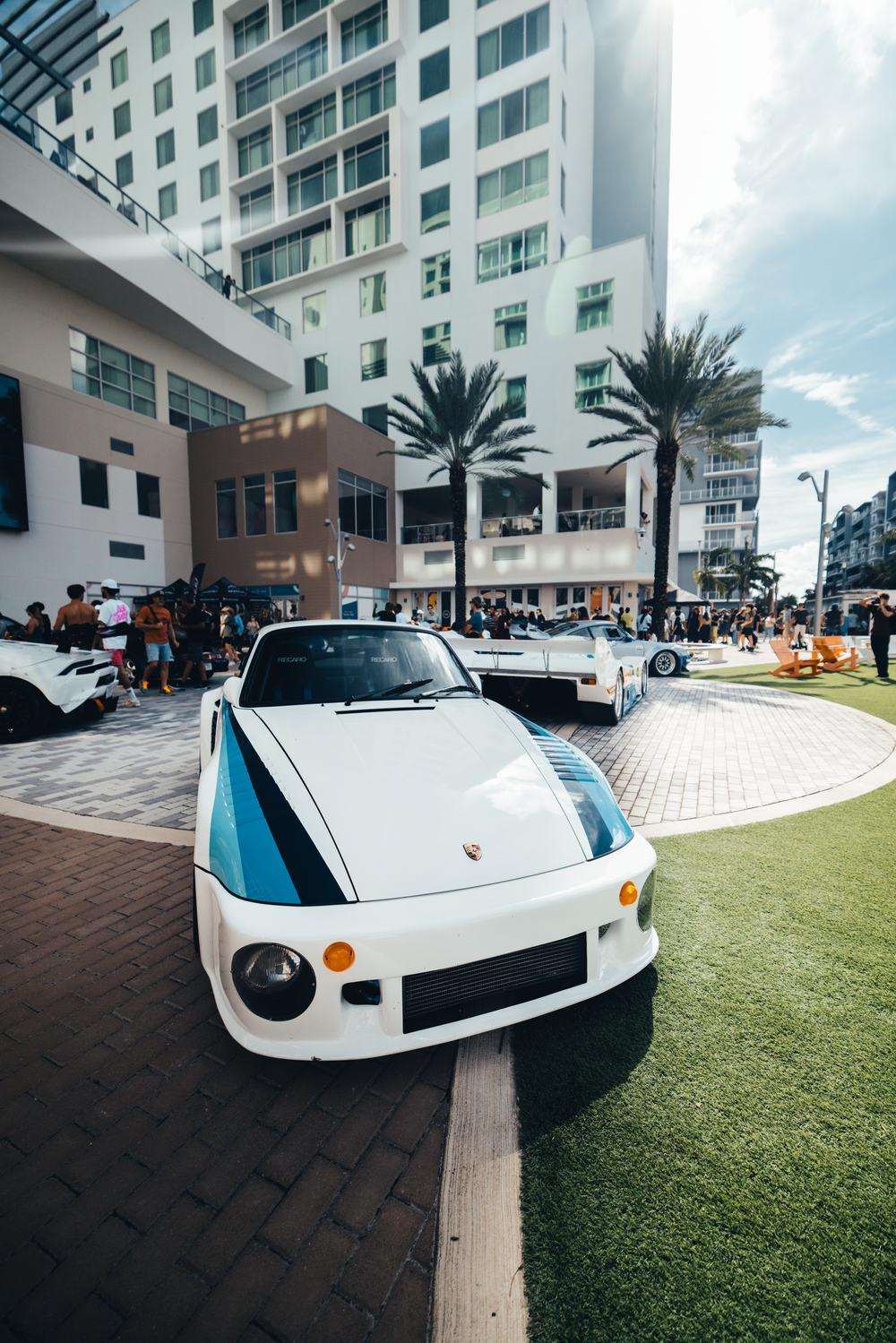 A white Porsche sports car with blue racing stripes parked in front of a modern building with palm trees.