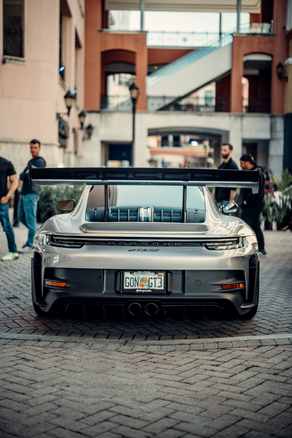Rear view of a silver Porsche GT3 sports car parked on cobblestone street with distinctive tail lights and exhaust.