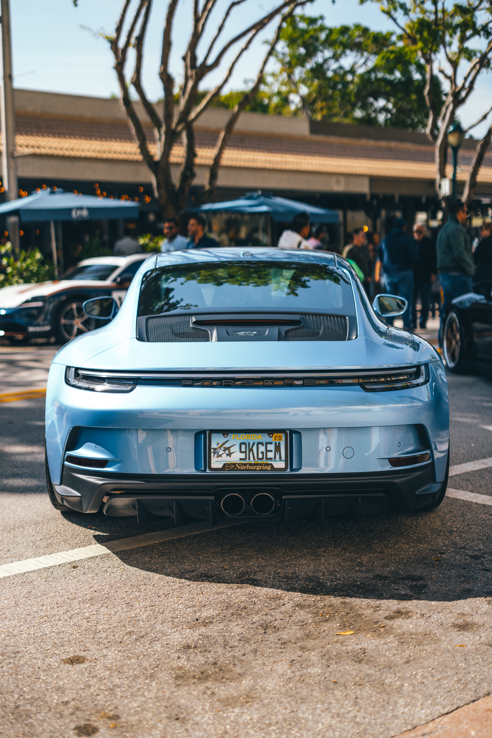 A sleek light blue Porsche 911 GT3 with dual exhaust tips parked in a shopping center parking lot.