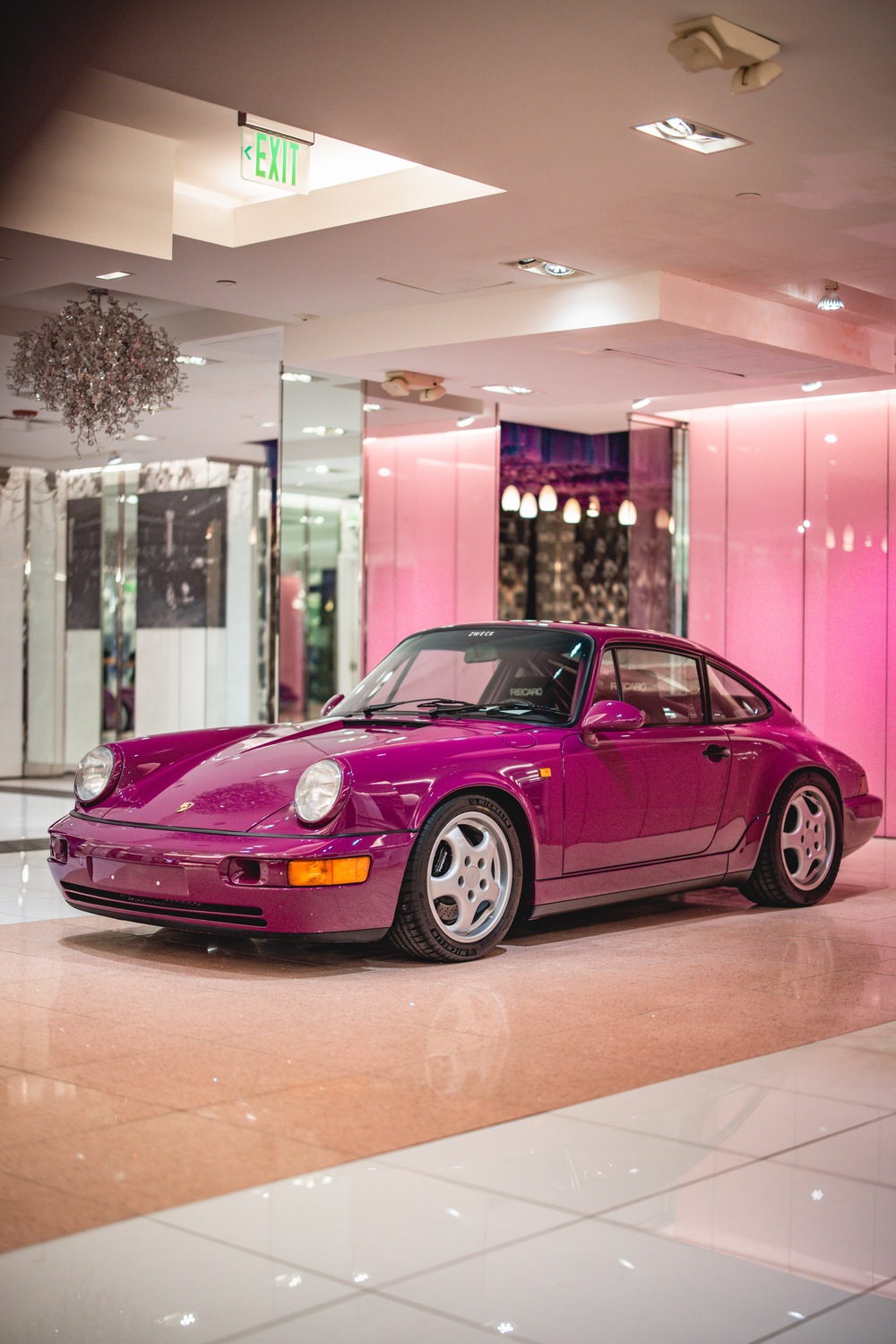 A stunning magenta Porsche 911 on display inside a modern mall with pink-lit walls and reflective floors.