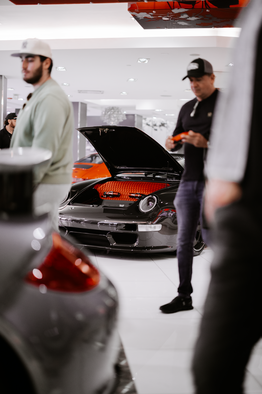 People examining a modified black sports car with exposed engine bay in indoor showroom space.