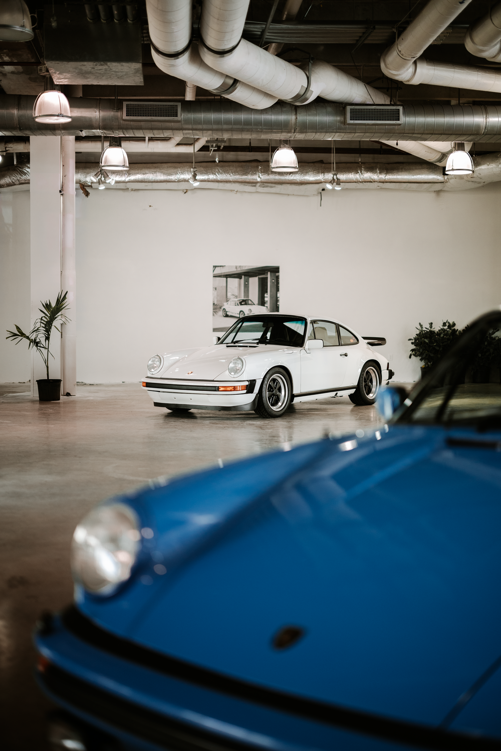 A classic white Porsche 911 and blue sports car in an indoor garage space with industrial ceiling details.