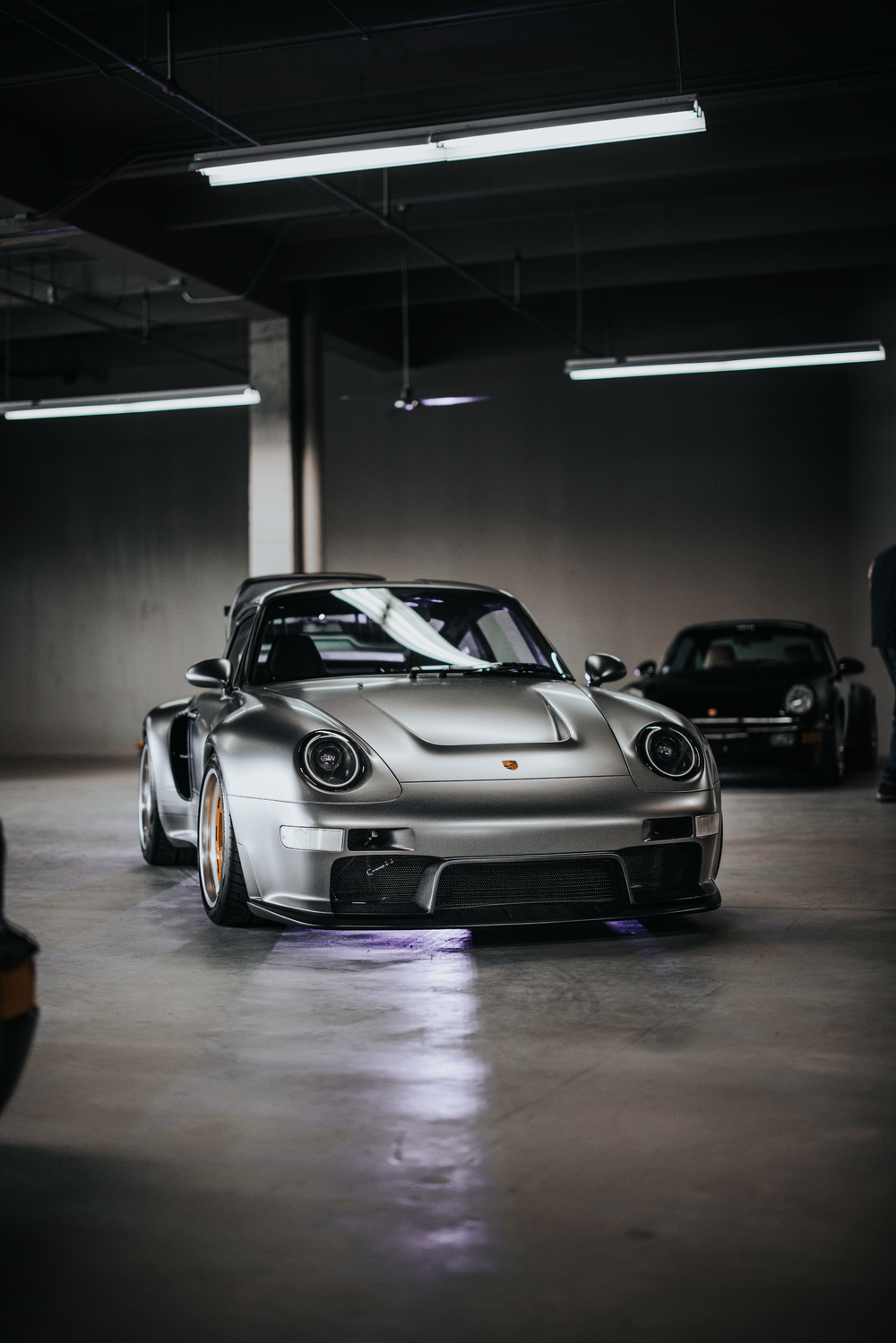 A silver Porsche 911 with custom modifications sits in a dimly lit garage under fluorescent lighting.
