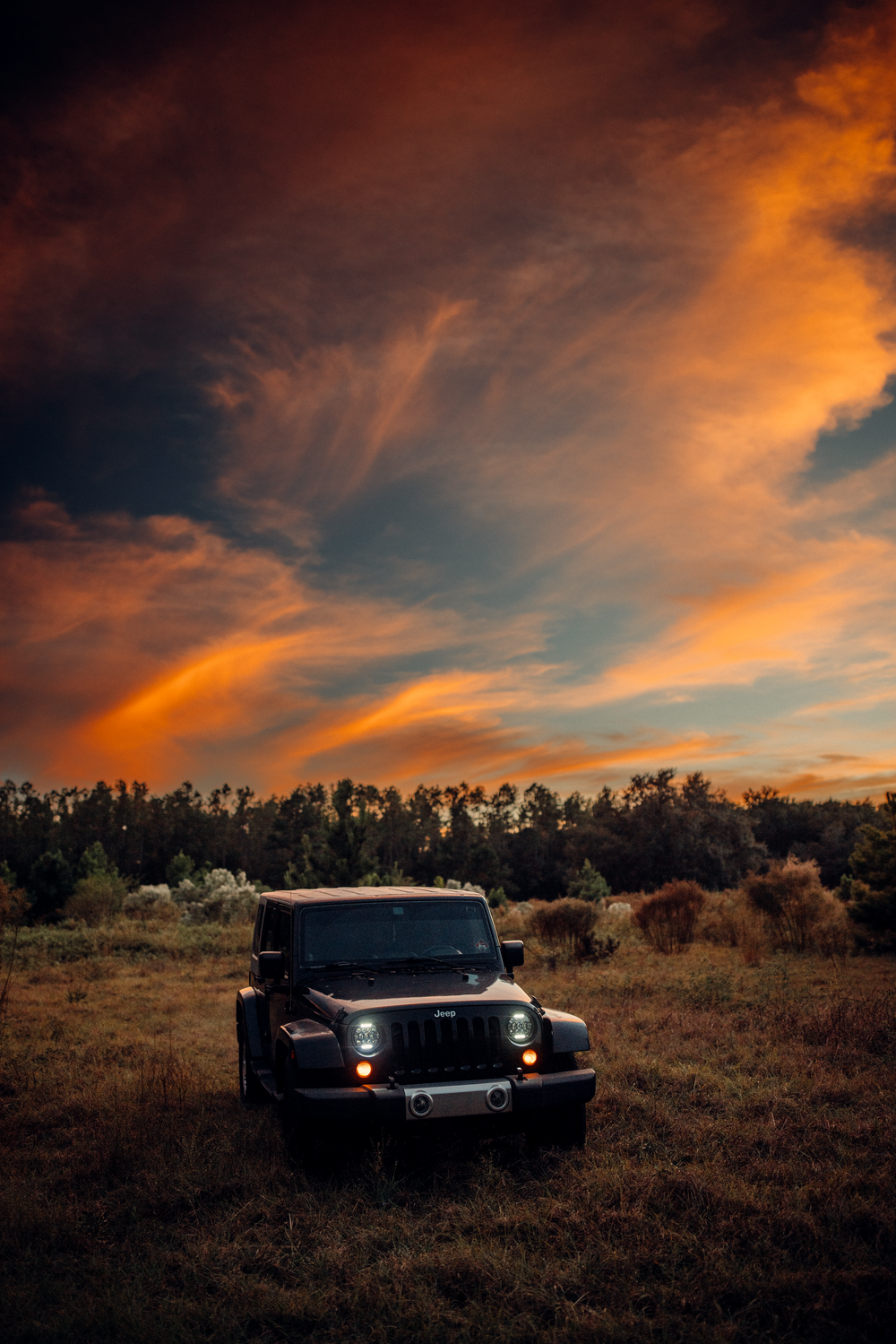 A black Jeep Wrangler parked in a field during dramatic sunset with orange clouds and dark blue sky.