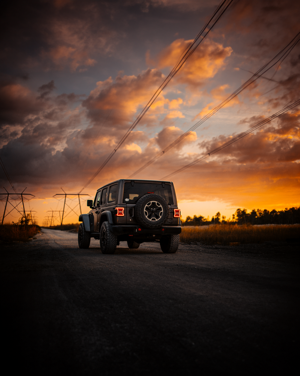 A black Jeep Wrangler silhouetted against a dramatic sunset sky with orange clouds and power lines overhead.