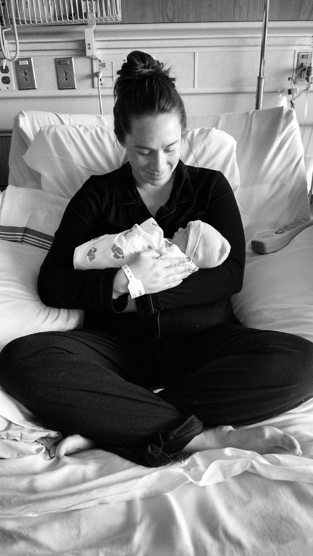Black and white photo of a woman in dark clothing holding a newborn in a hospital bed.