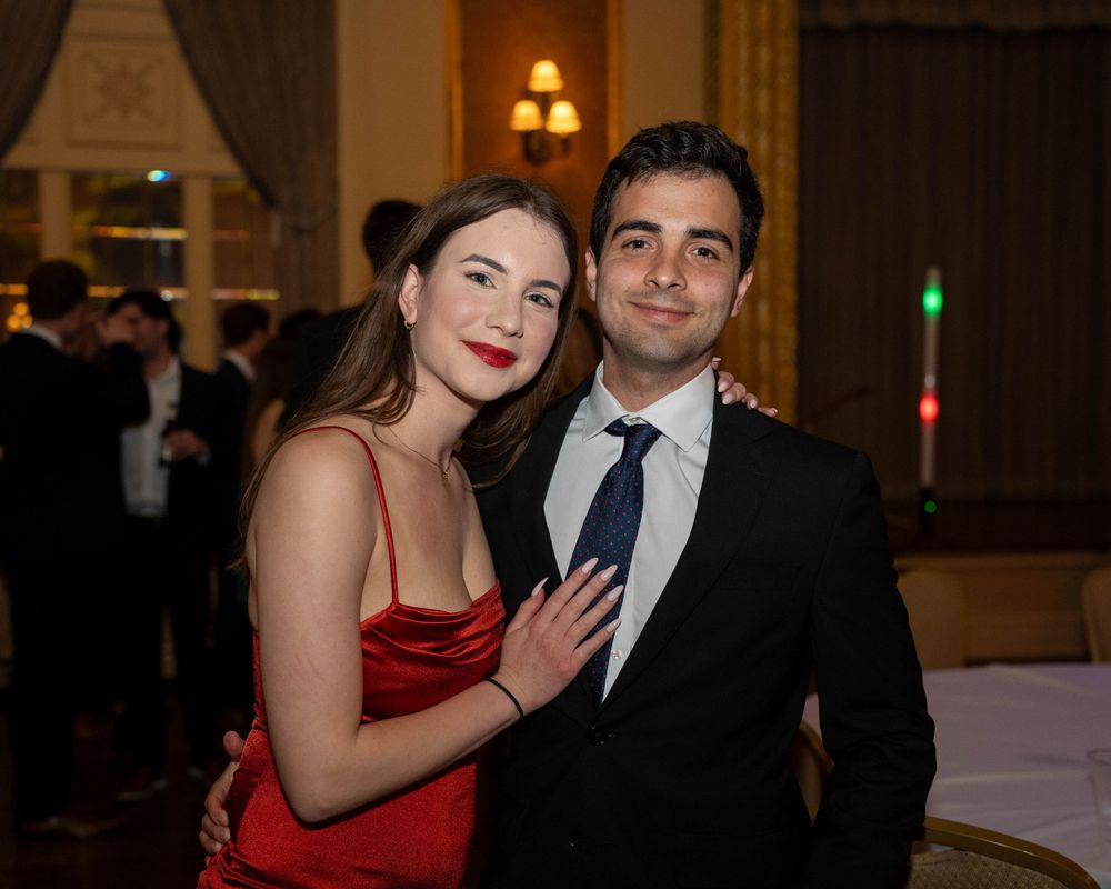 A couple poses together at a formal event, with one wearing a red satin dress and the other in a dark suit and tie.