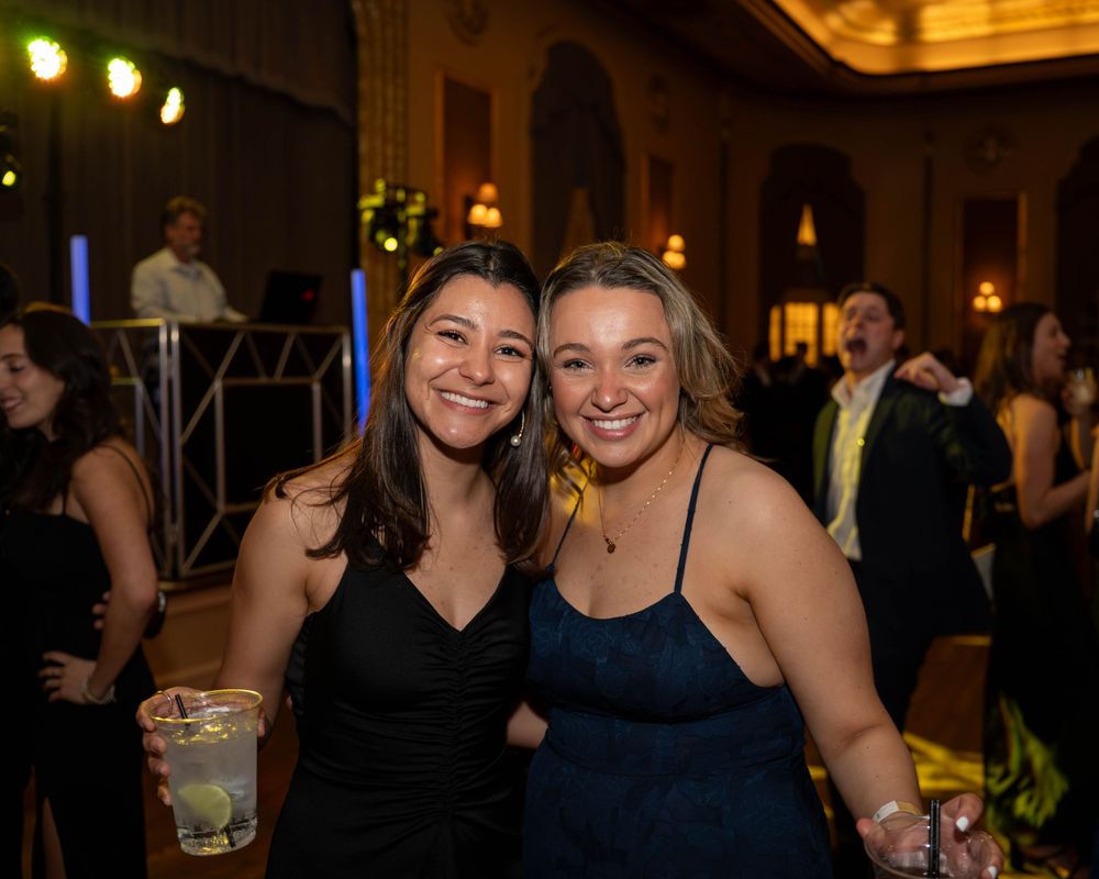 Two friends wearing black and navy dresses pose together at a festive indoor celebration.
