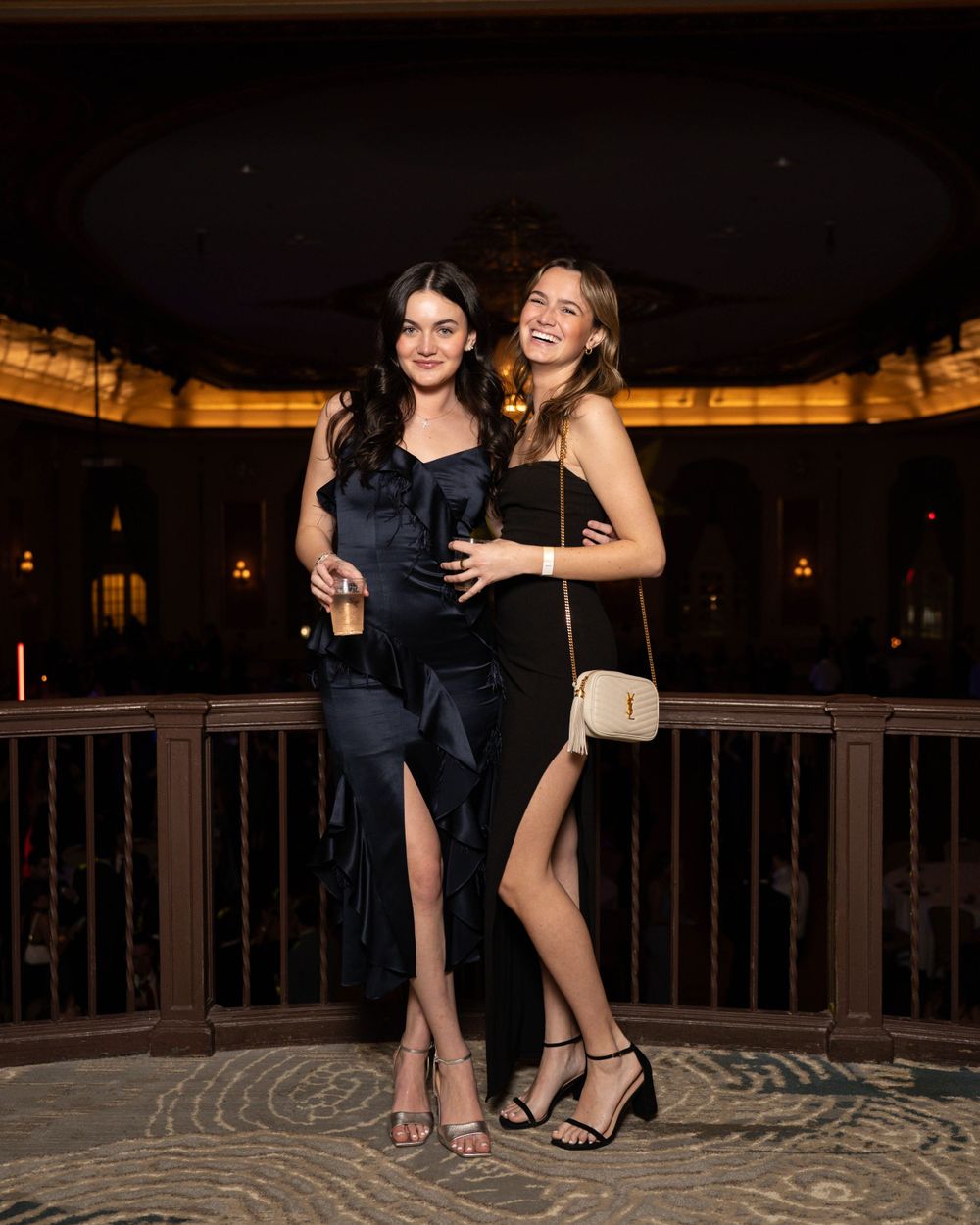 Two friends pose together in formal attire at an elegant indoor venue with decorative railings and mood lighting.