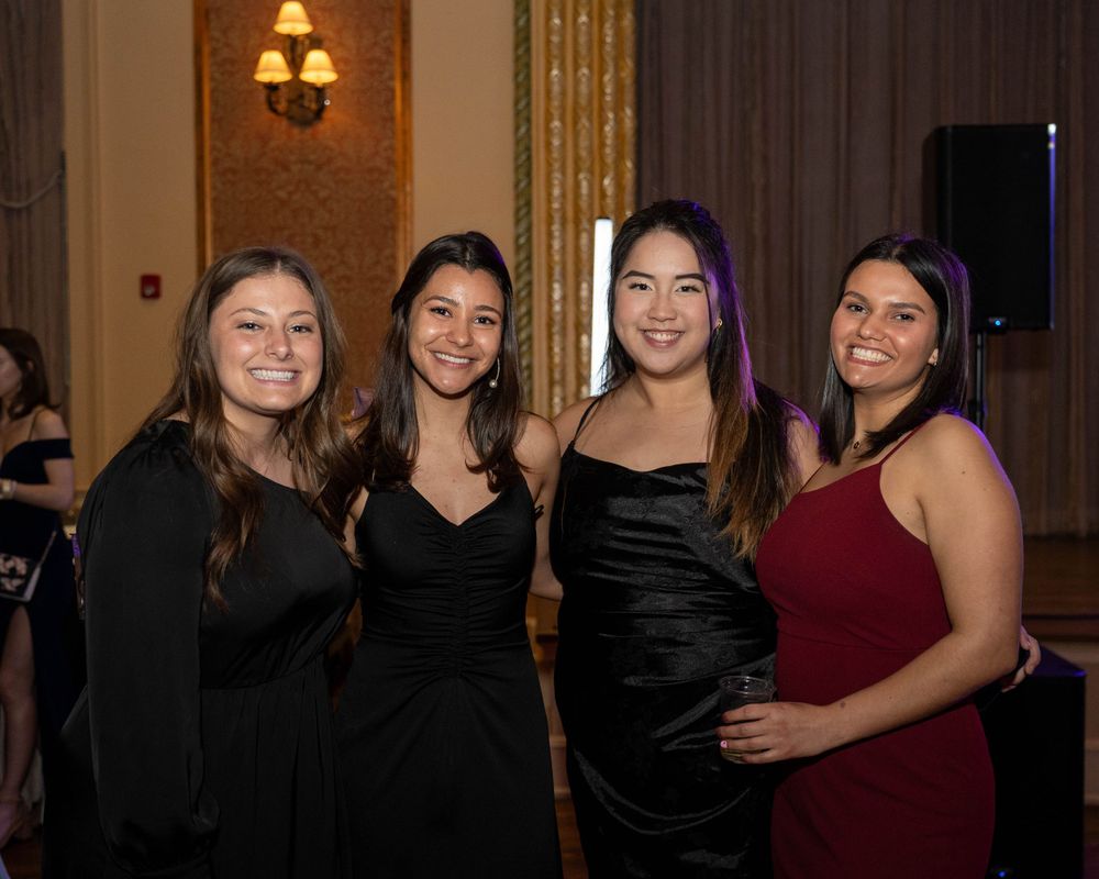 Four friends pose together at a formal event in a banquet hall with warm lighting and elegant decor.