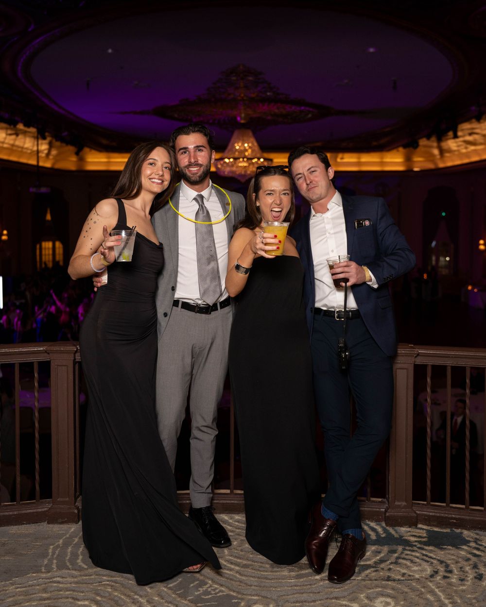 A group of four formally dressed attendees pose together at an elegant venue with ornate decor in the evening.
