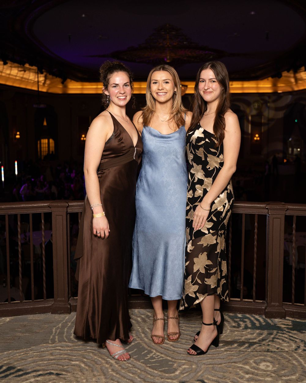 Three friends in formal attire pose together at an elegant venue with illuminated architecture in the background.