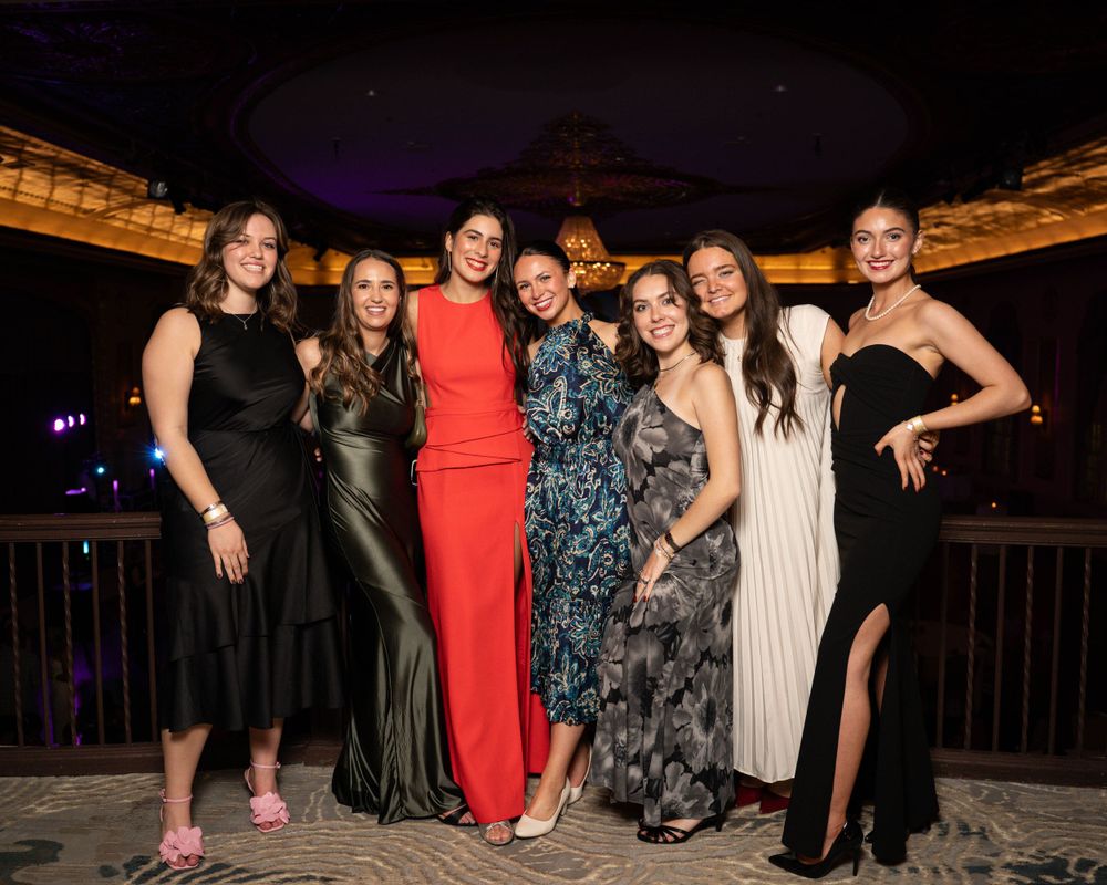 Group of friends dressed in elegant formal attire pose together at an evening event on a balcony with city lights behind them.