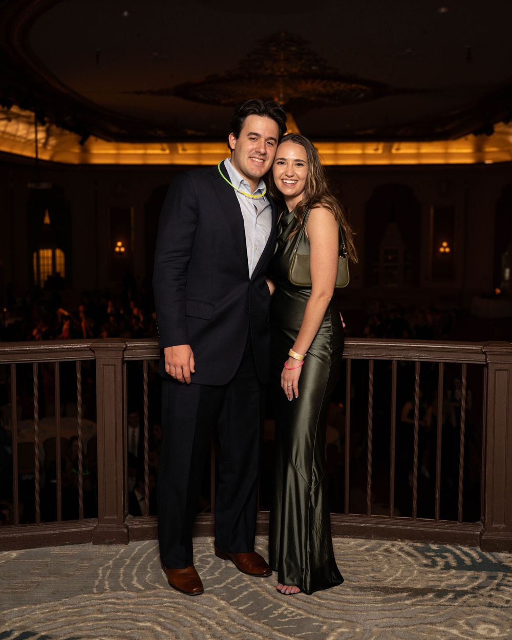 A couple poses together at an elegant venue, with the woman wearing a long olive green dress and the man in a dark suit.