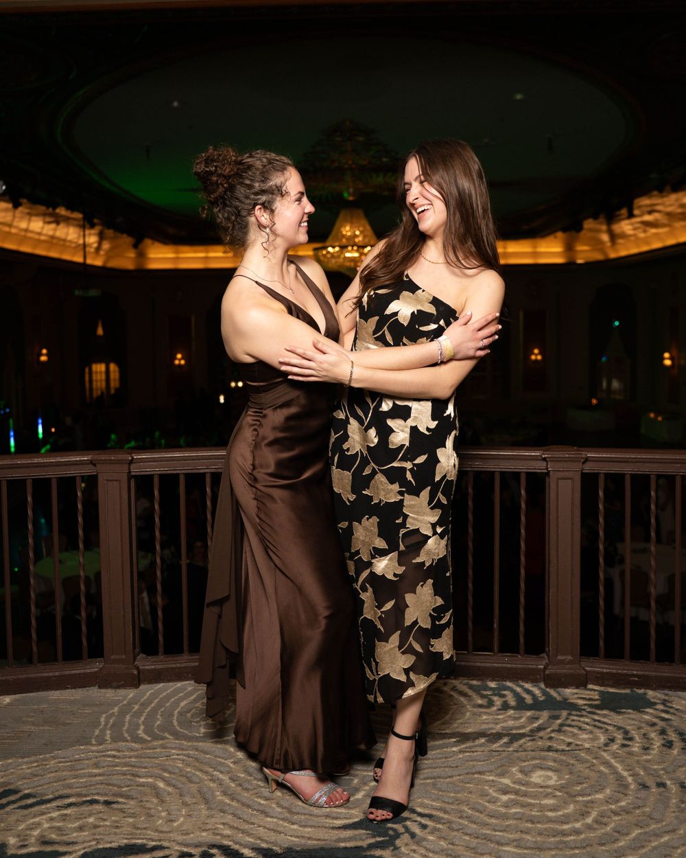 Two friends sharing a joyful moment together while wearing formal dresses at an evening event.
