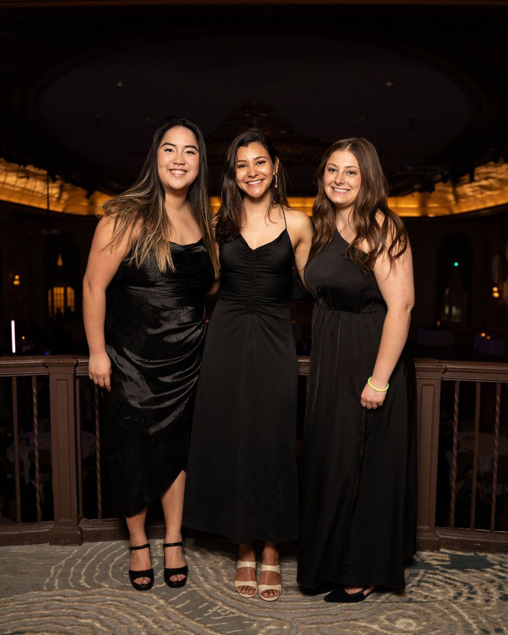 Three women in elegant black dresses pose together at a formal evening event on a wooden deck with warm lighting.