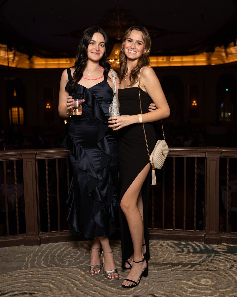 Two women in black formal dresses pose together on balcony at nighttime event.
