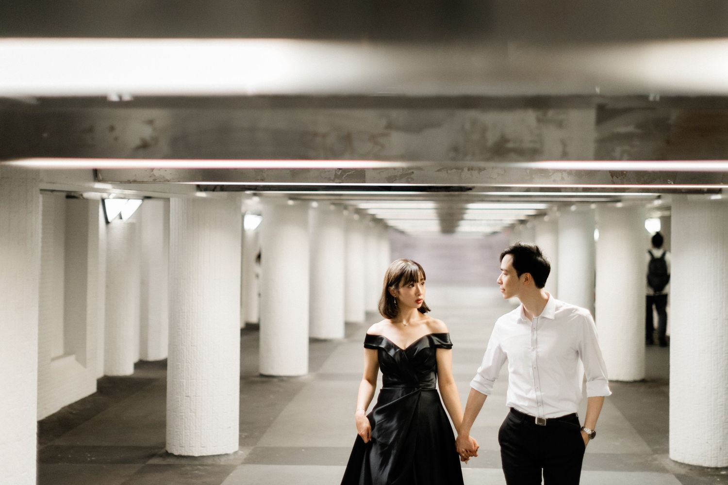 Smiling couple in formal attire holding hands walking through a pillared underground parking structure.