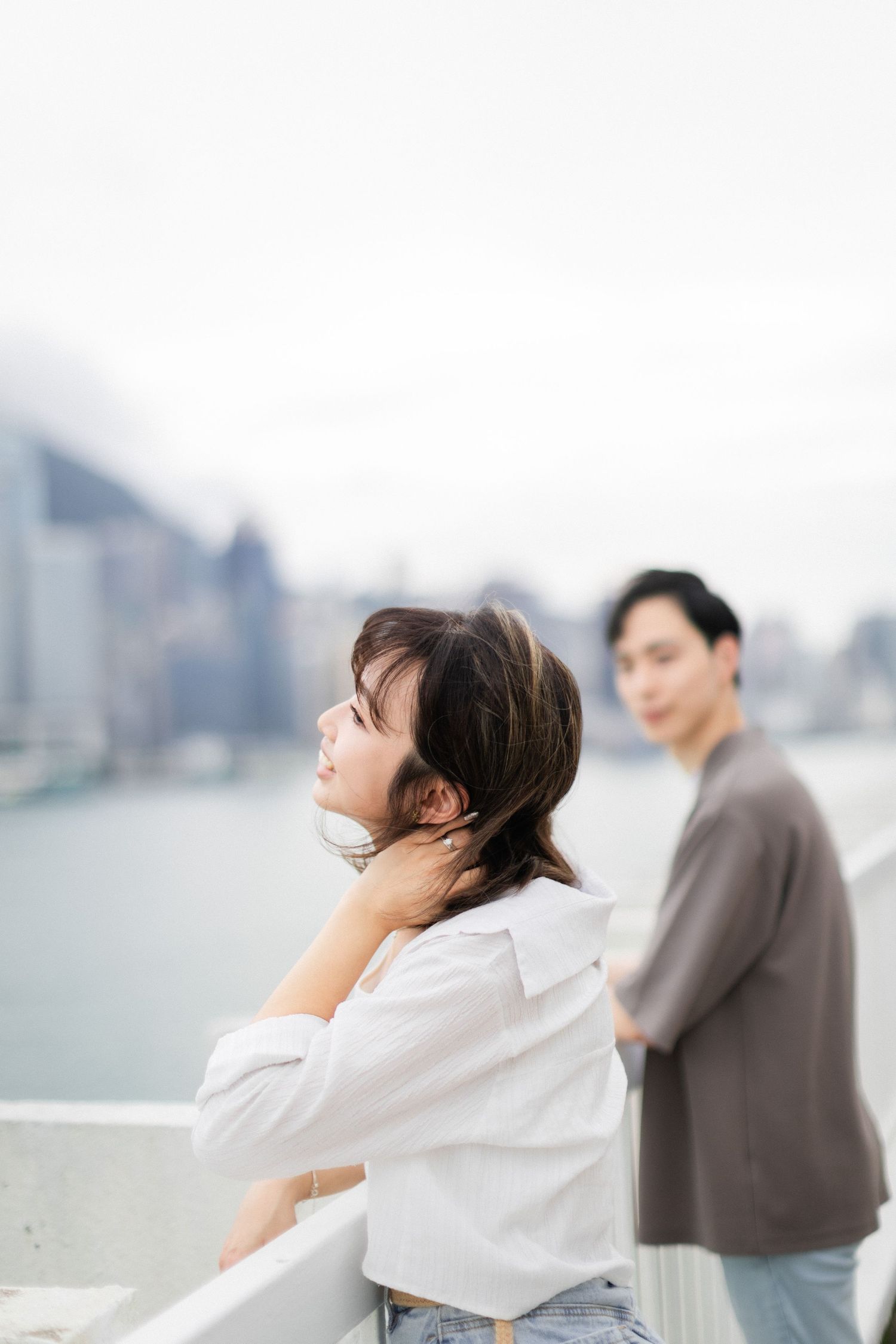 Young woman in white shirt leans on railing gazing at Hong Kong harbour skyline while man stands behind her.