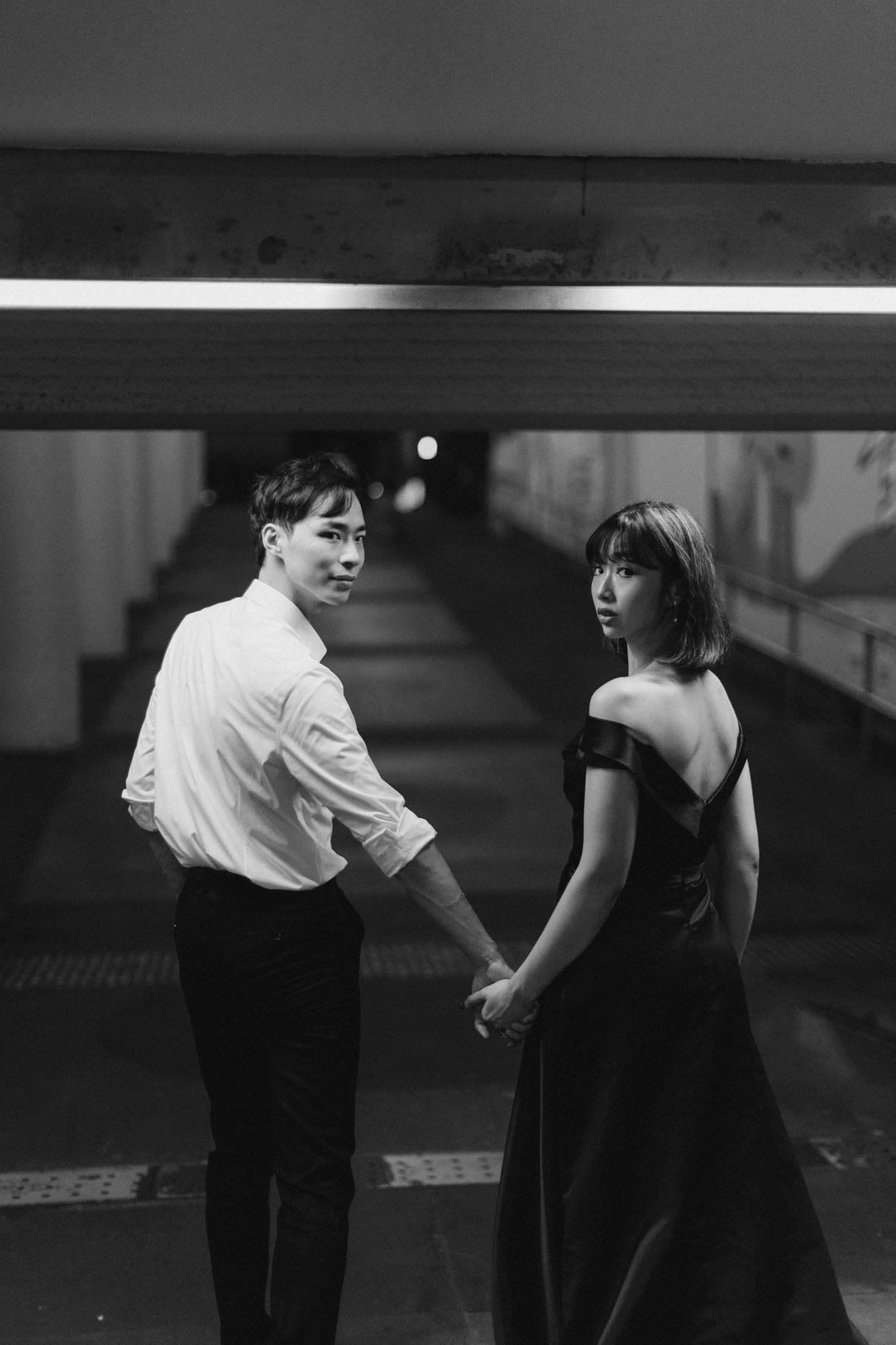 Black and white romantic photo of a couple holding hands and glancing back in an urban underpass.