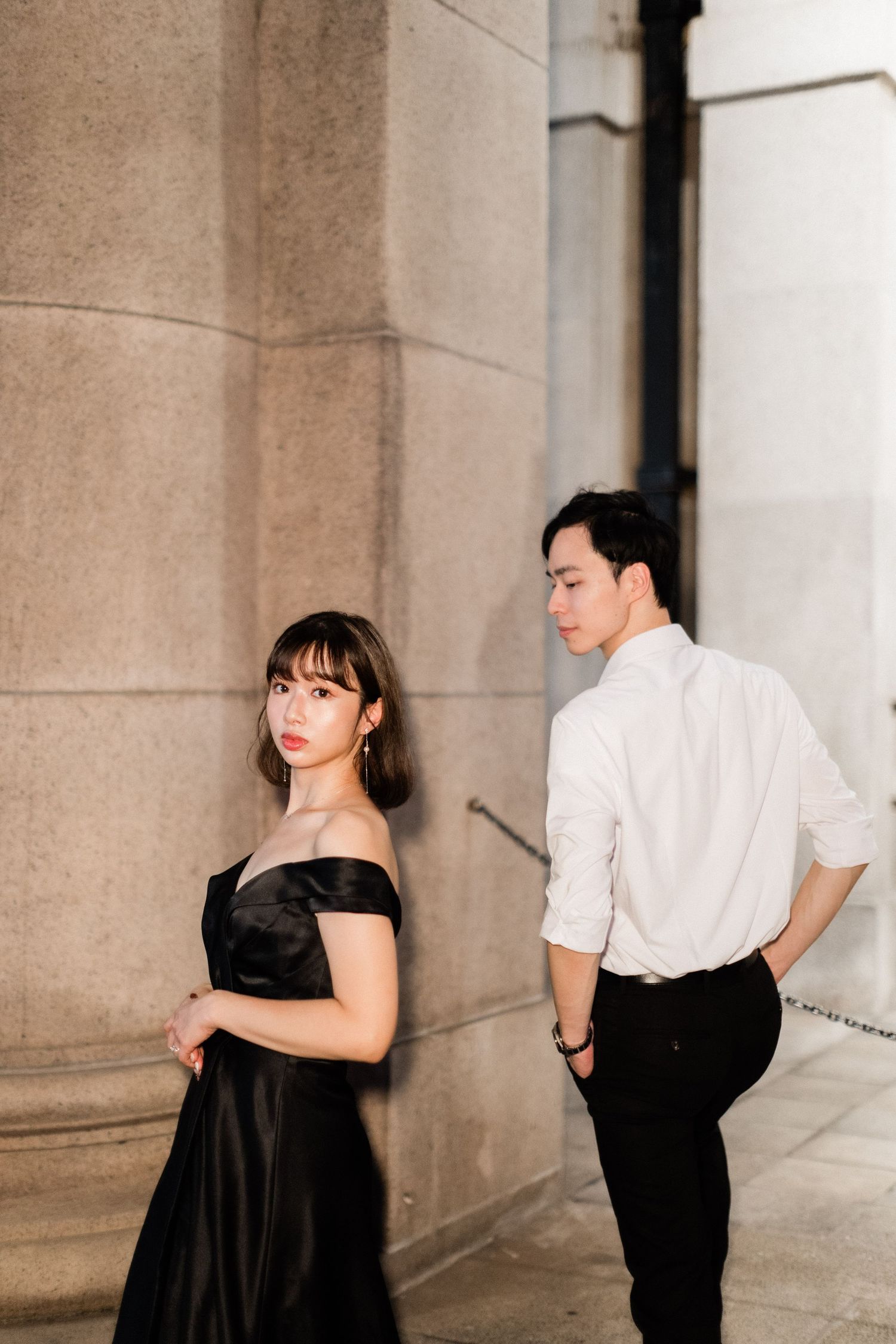 Couple posing by stone column, woman in black off-shoulder gown and man in white shirt at night.