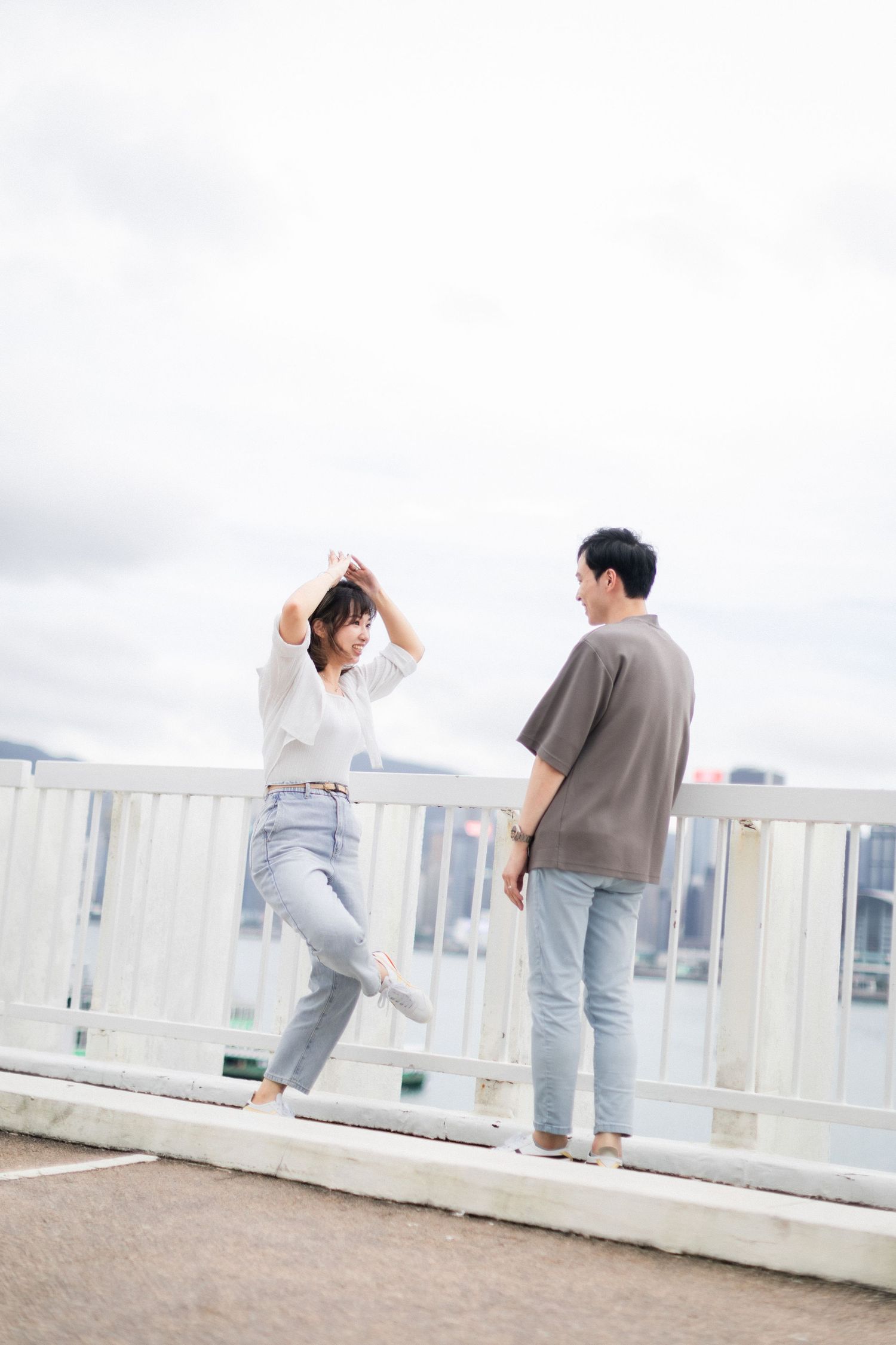 Playful couple dancing together on waterfront promenade with Hong Kong city skyline in background.