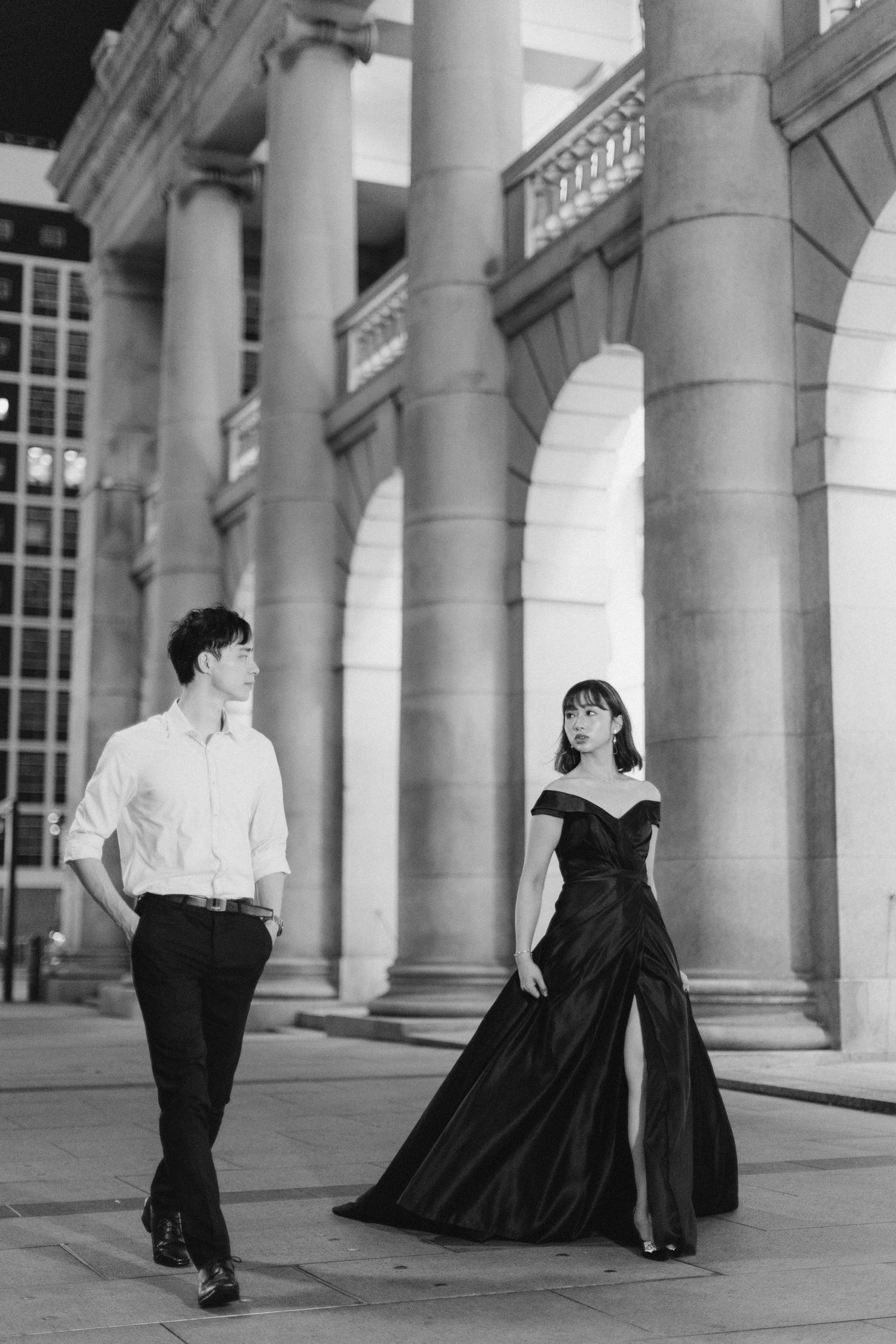 Elegant couple walking apart near classical stone columns, woman in dramatic black gown, black and white photo.