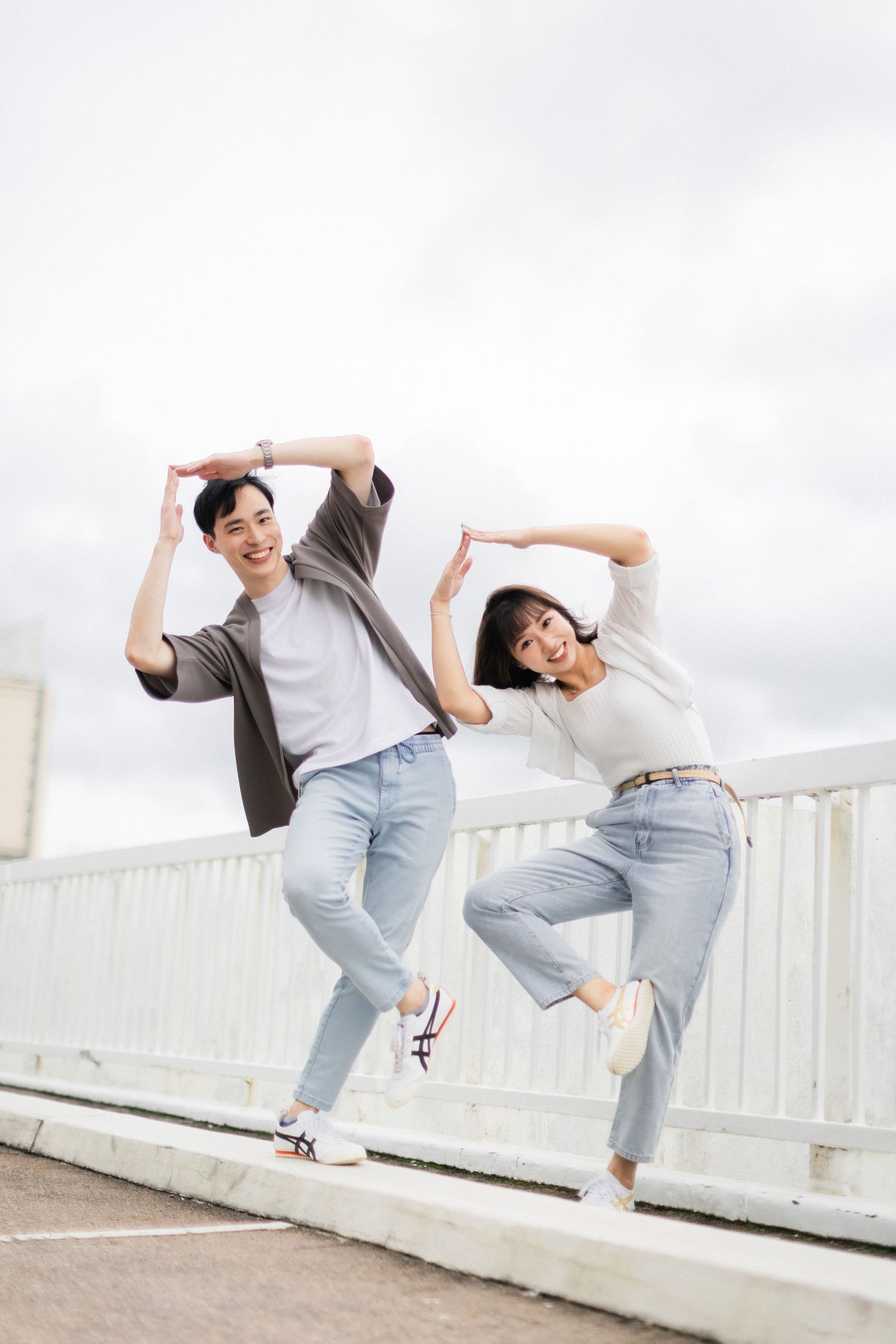 Joyful Asian couple jumping and posing together on rooftop with cloudy sky behind them.