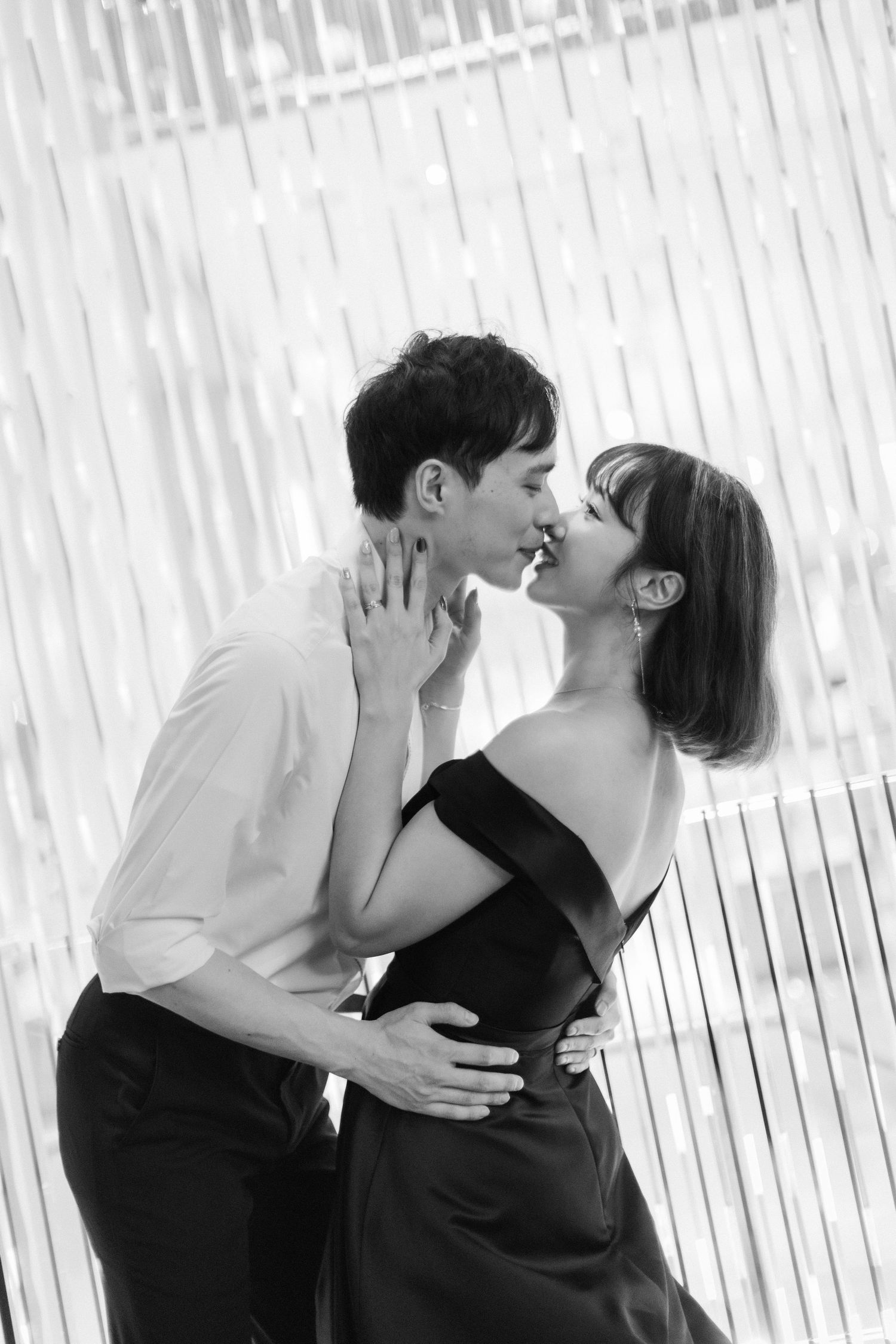 Romantic couple sharing a kiss, woman in black off-shoulder dress, intimate black and white portrait.