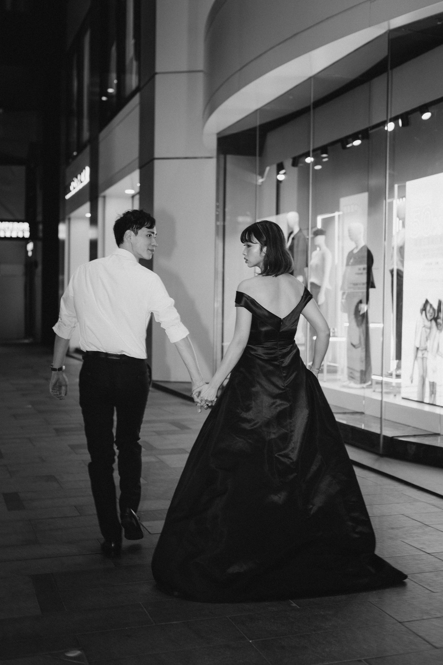 Couple holding hands walking past luxury store at night, woman in flowing black gown, black and white photo.