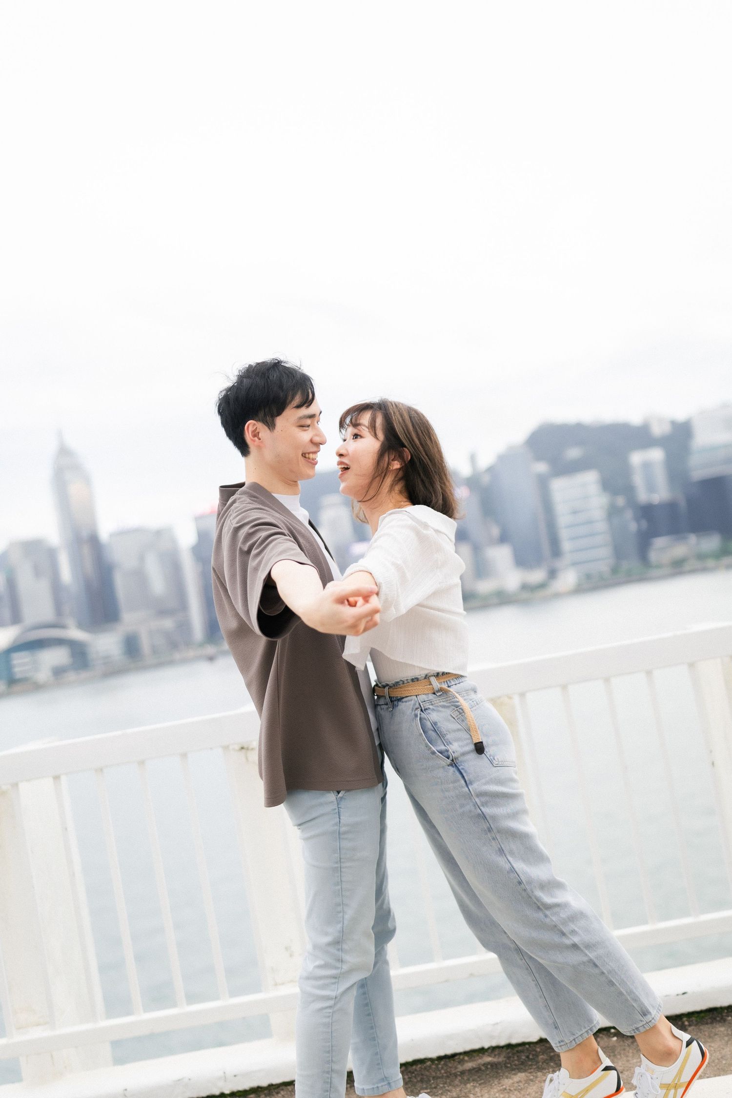 Happy Asian couple holding hands and laughing together on waterfront with city skyline backdrop.