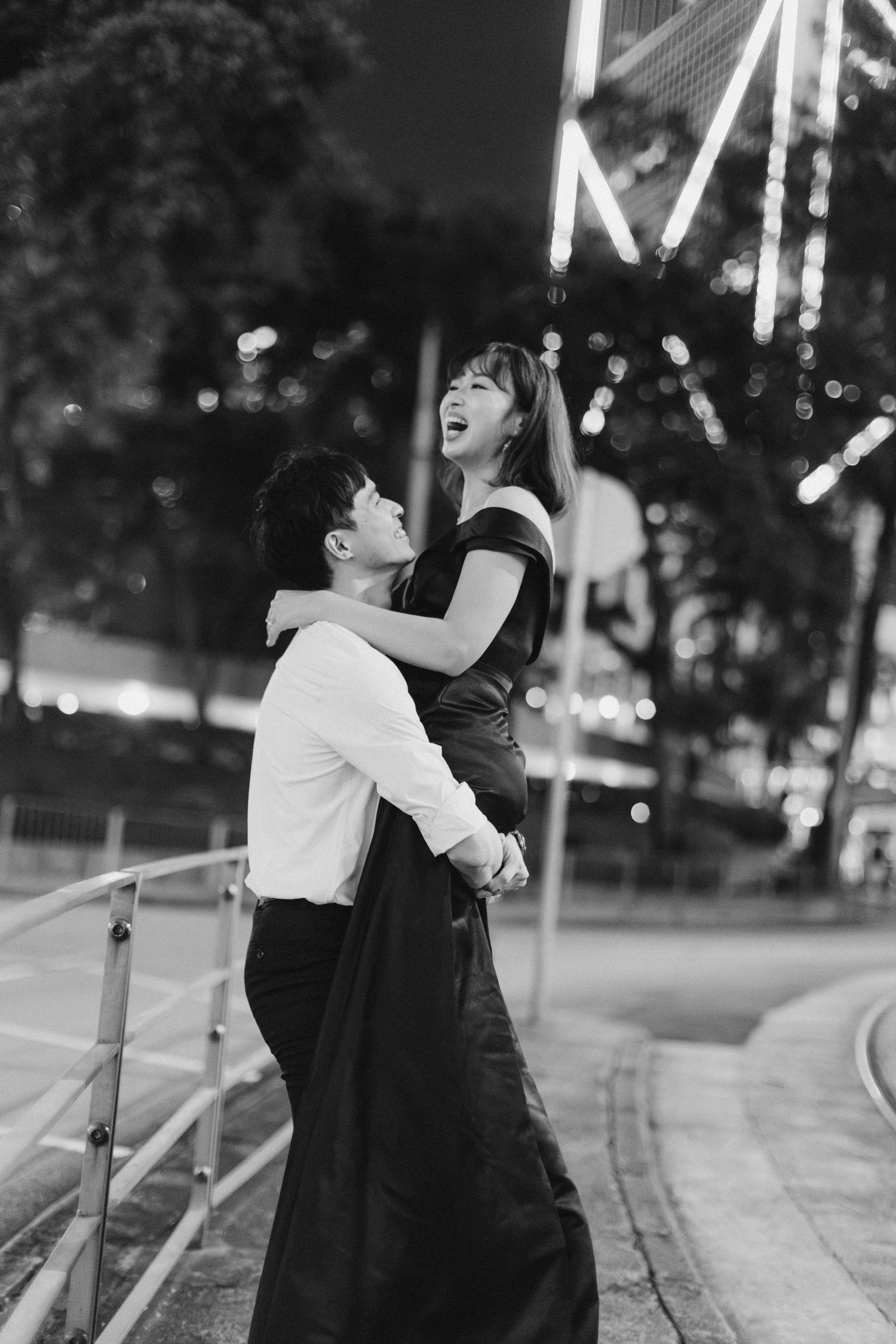 Couple embracing outdoors at night, woman laughing joyfully while being lifted, black and white portrait.