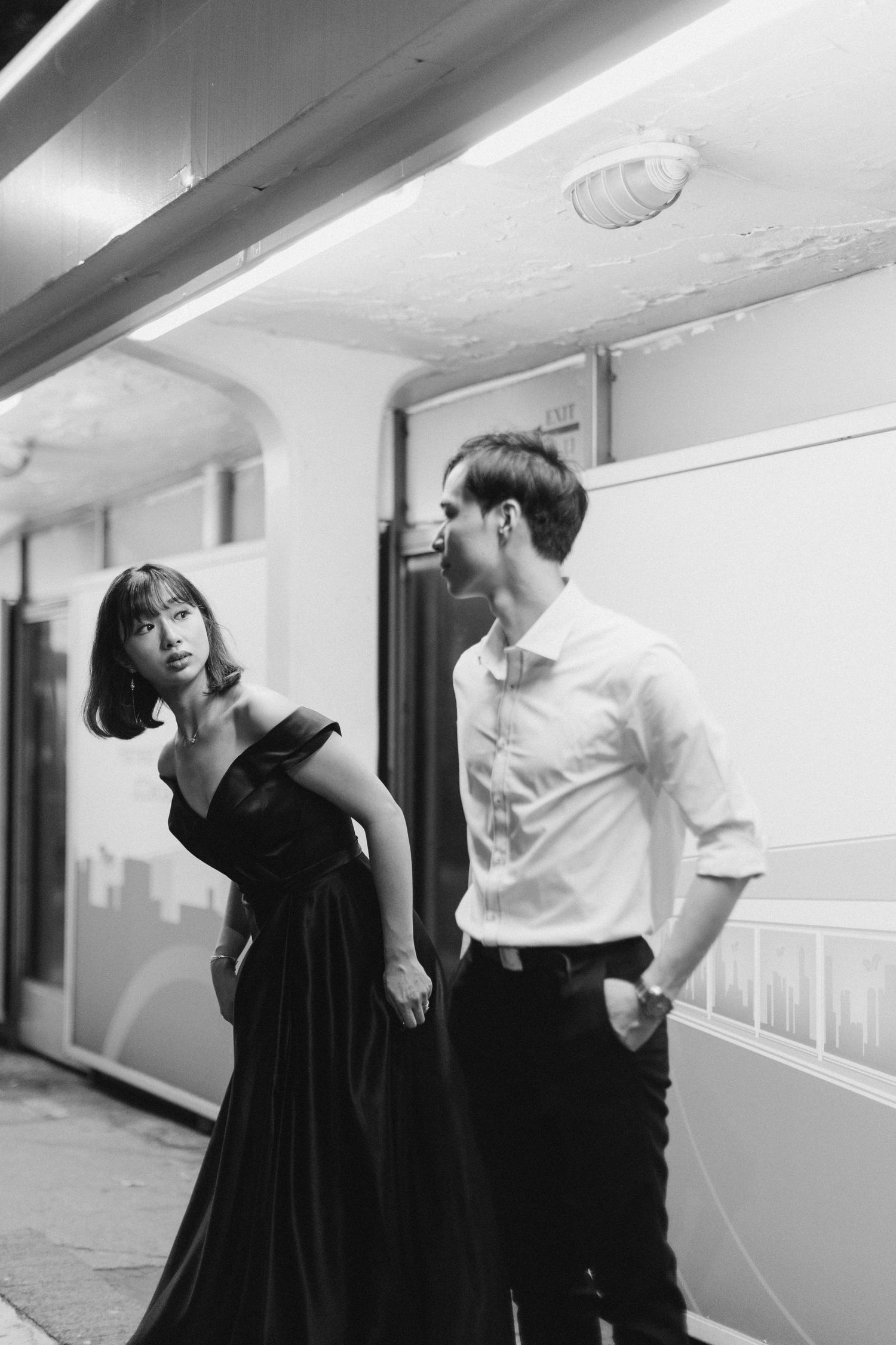 Couple holding hands in urban alleyway, woman in black gown glancing back, moody black and white photo.