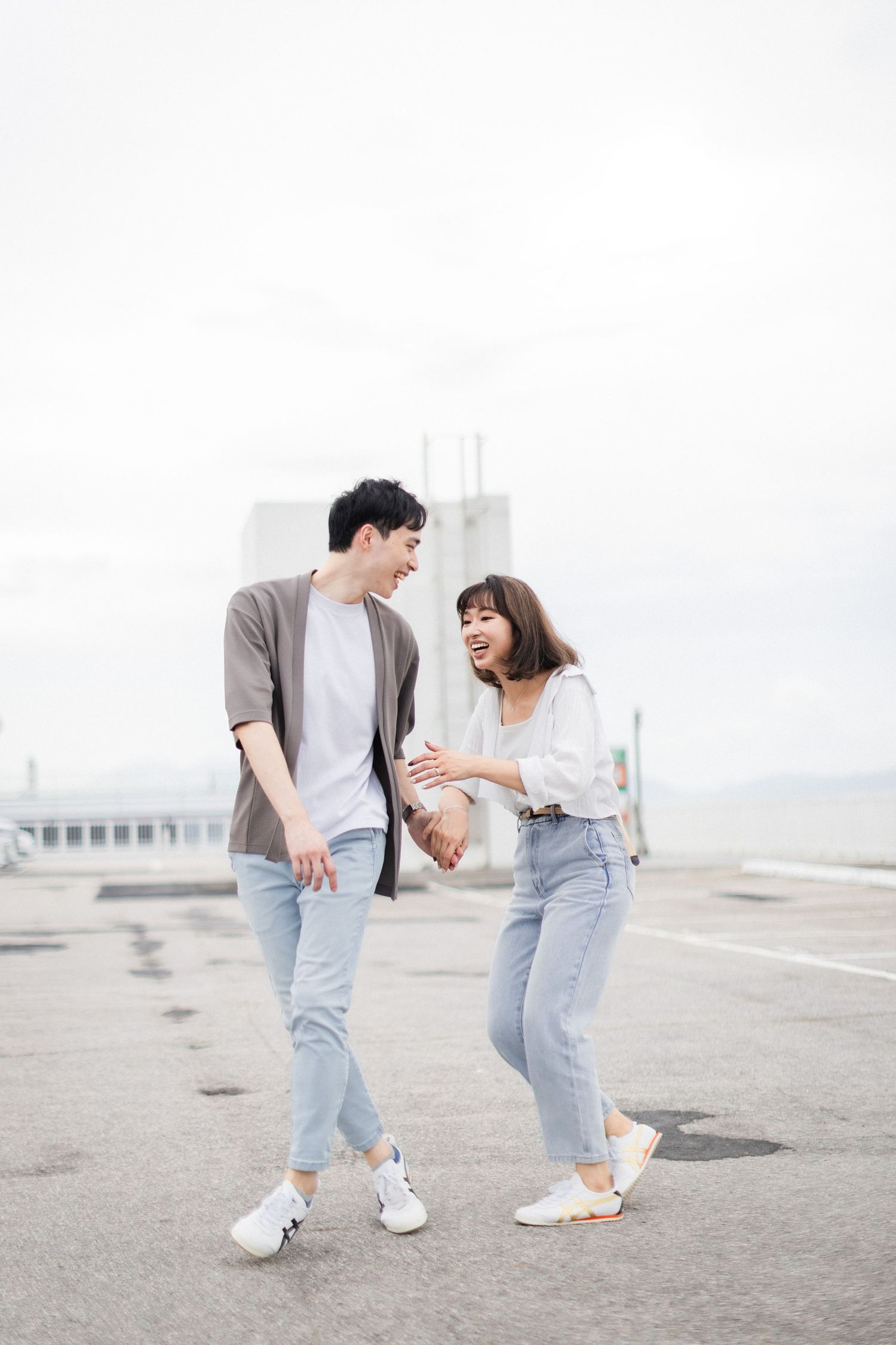 Smiling Asian couple walking hand in hand on rooftop while laughing together on overcast day.