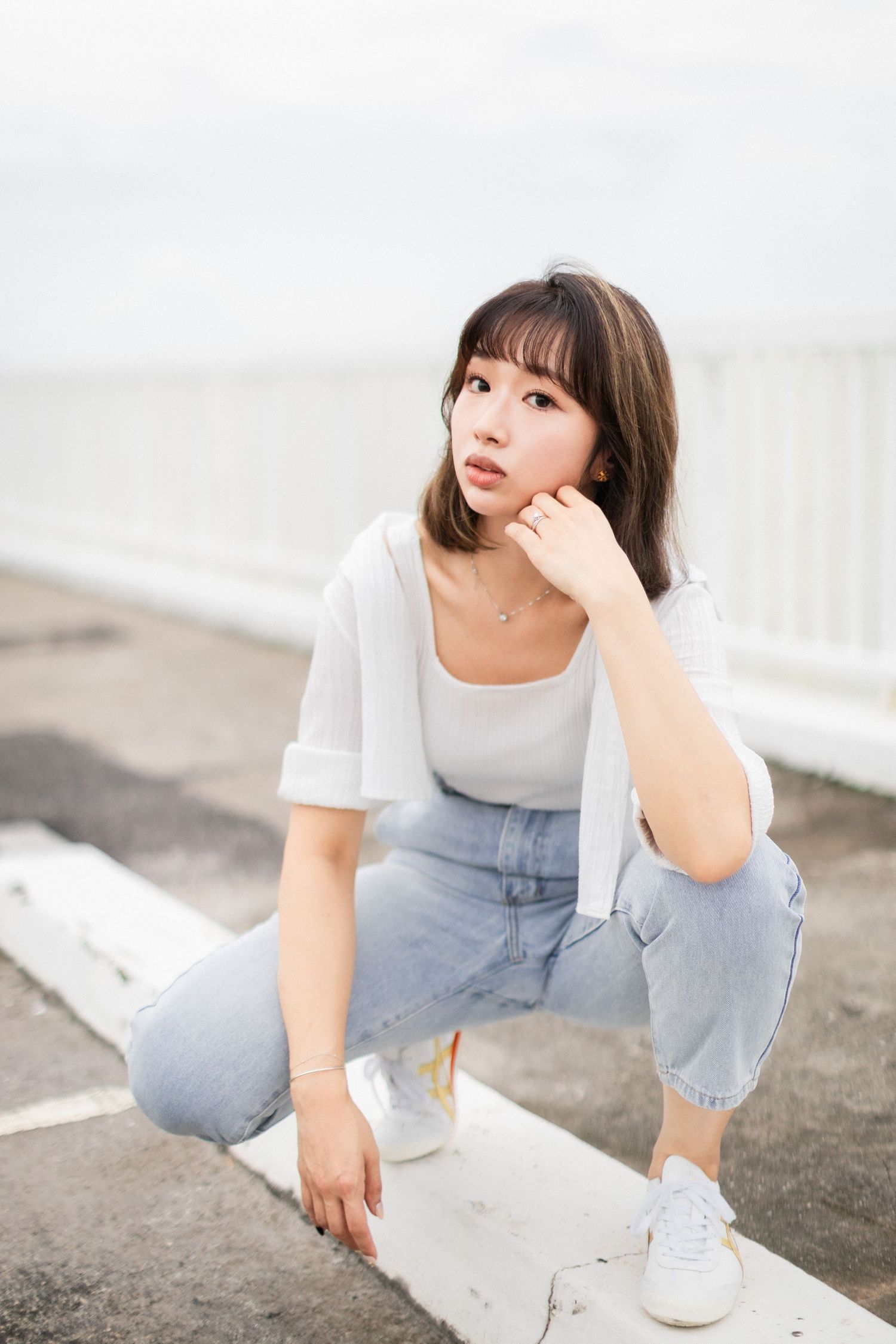 Young Asian woman in white top and jeans crouching thoughtfully on outdoor pavement near waterfront.