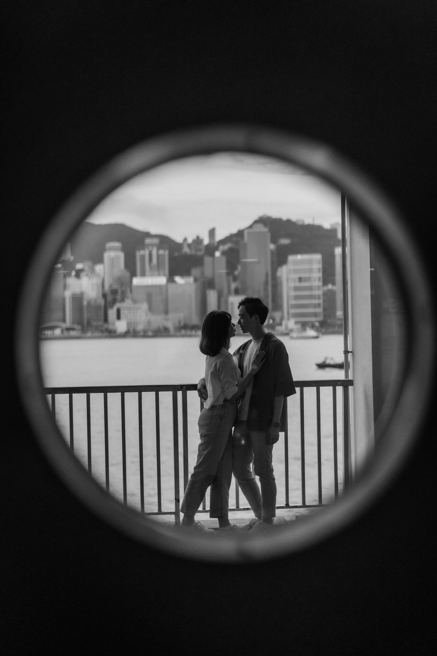 Romantic couple silhouetted through circular window with Hong Kong Victoria Harbour skyline in black and white.