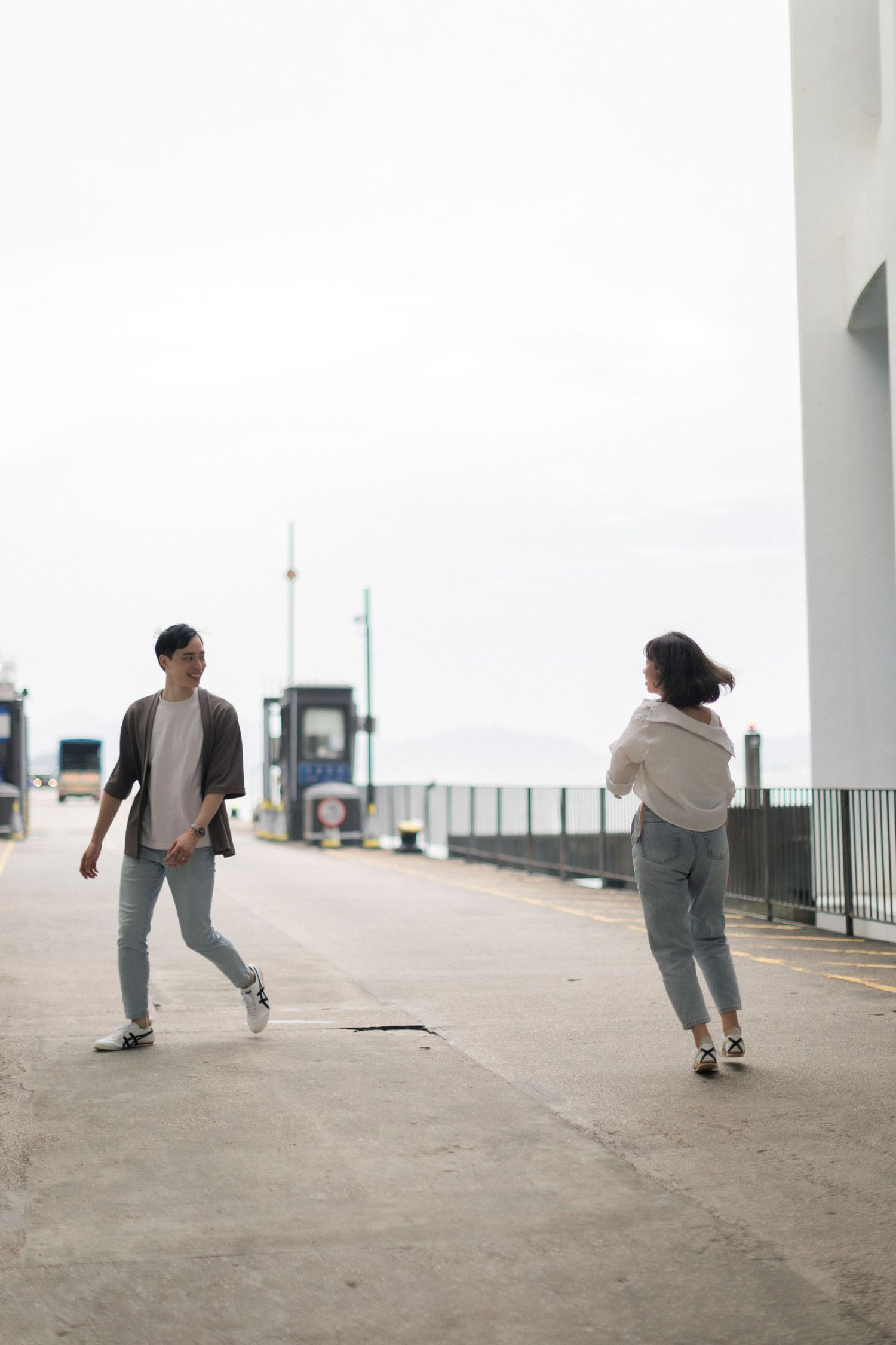 Young Asian couple walking toward each other on open harbour pier with overcast sky above.