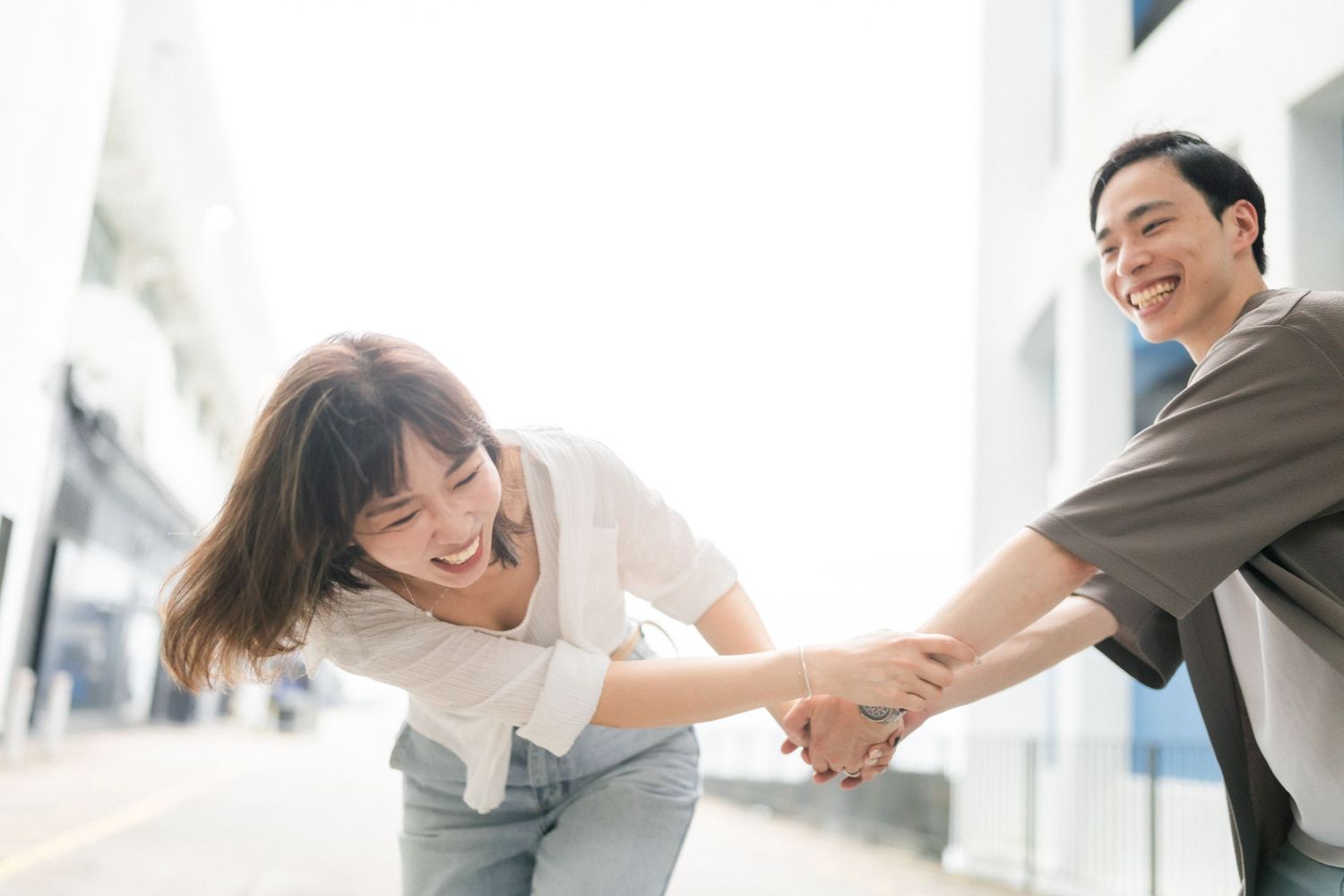 Laughing Asian couple holding hands and leaning forward together on bright sunny city street.