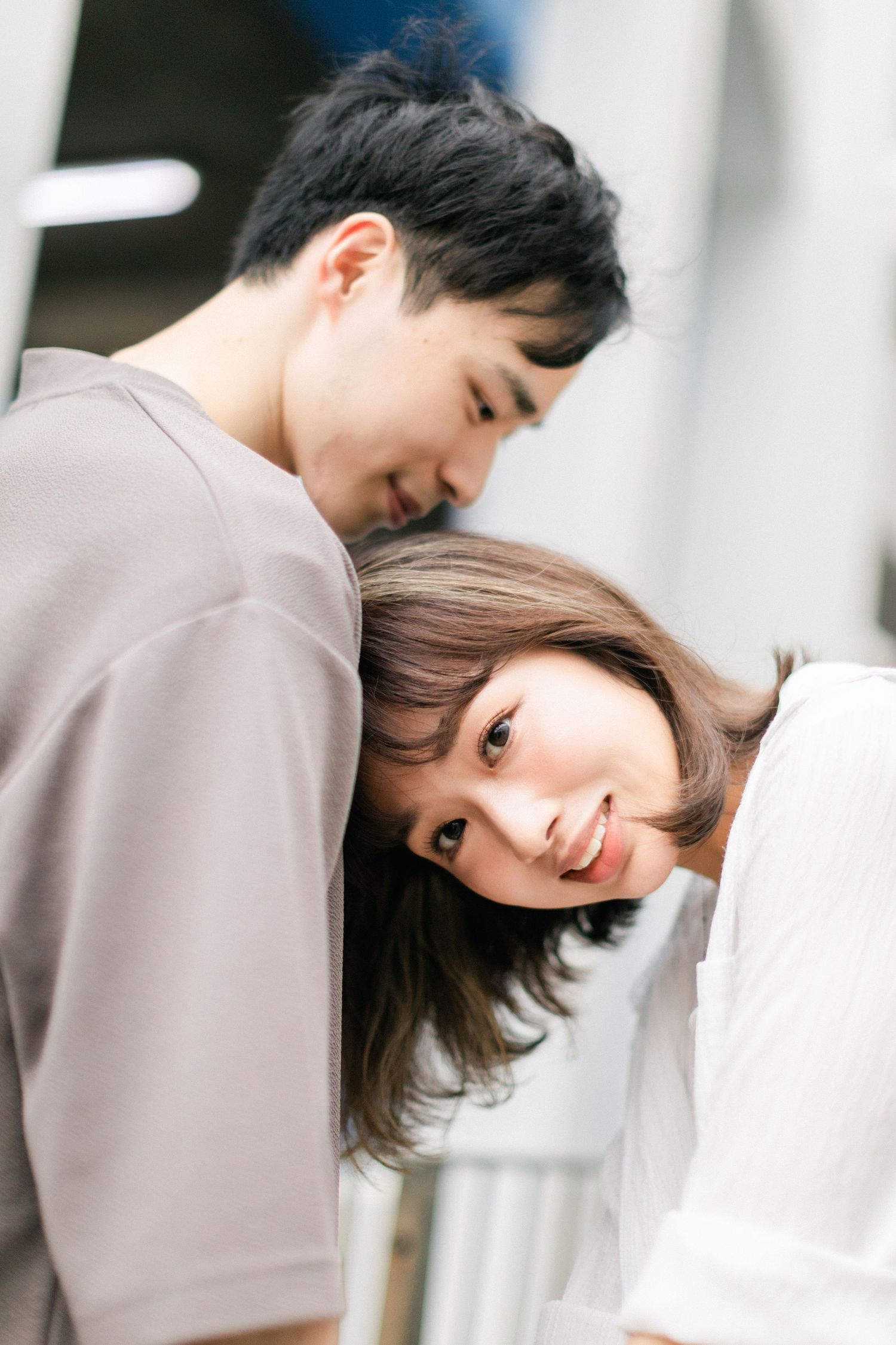 A young Asian couple sharing an intimate moment, woman resting her head on mans shoulder smiling at camera.