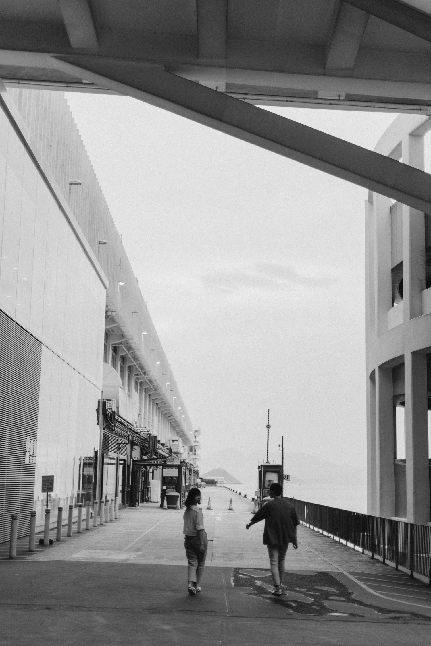 Black and white photo of a couple walking along a waterfront promenade between modern buildings.