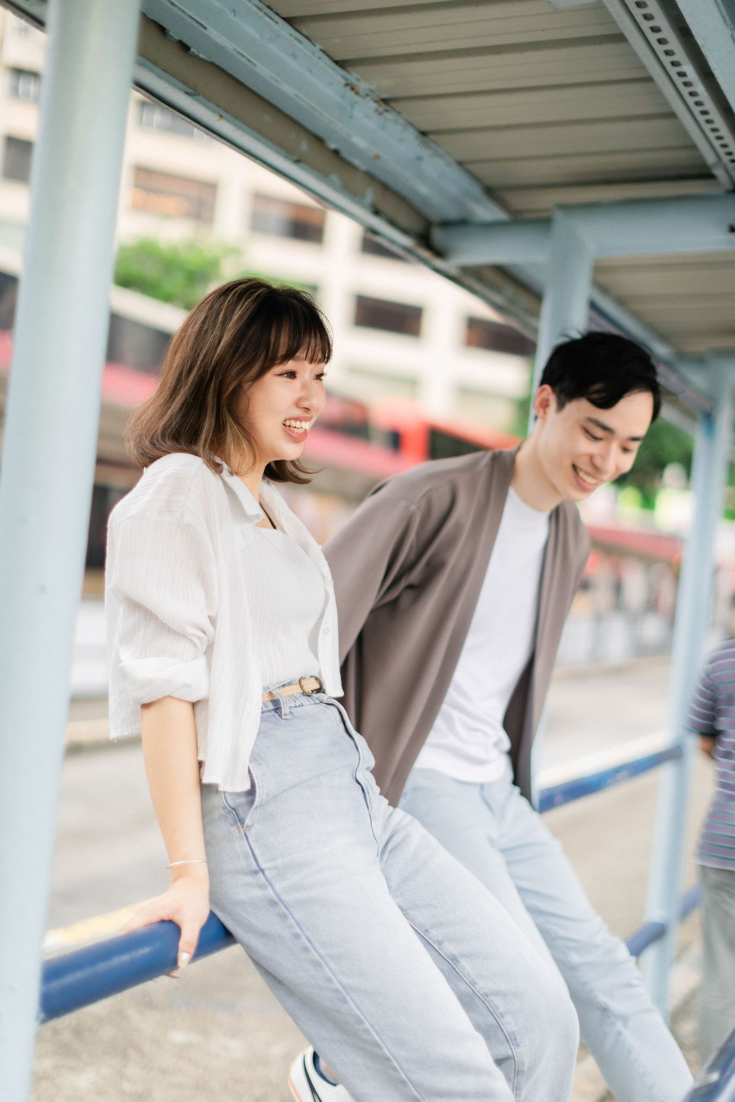 Happy Asian couple laughing and leaning against blue metal railings at a bus station outdoors.