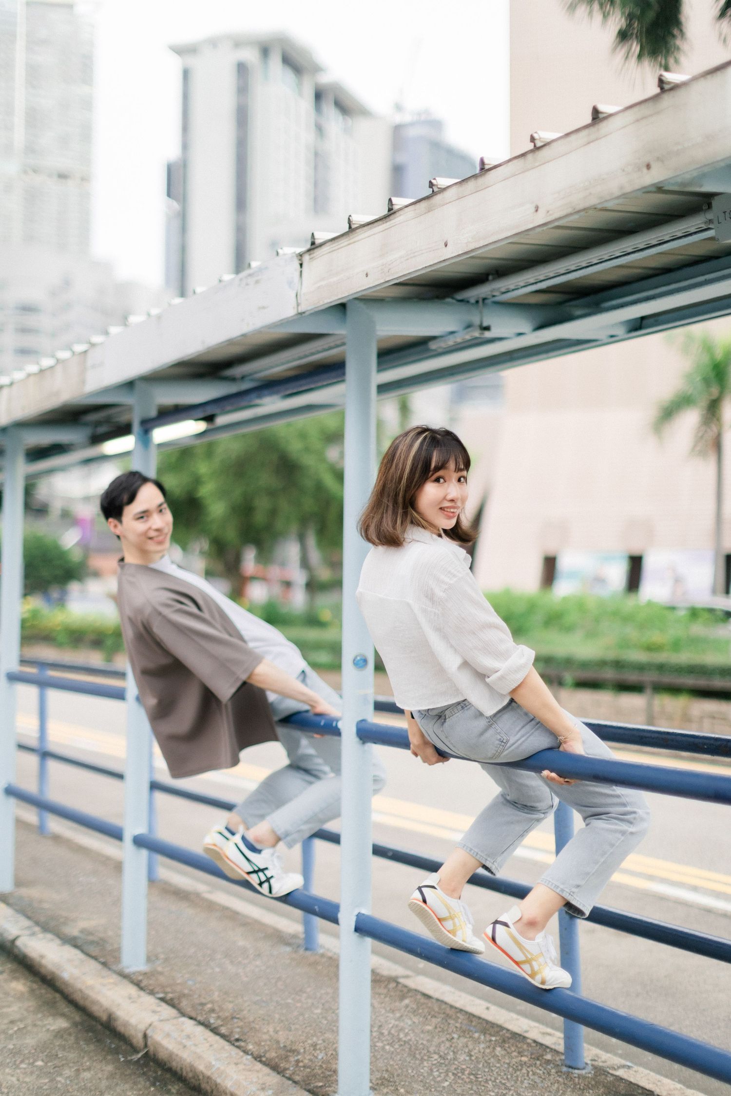 Two young Asian people posing playfully on blue railings at an urban bus stop with city buildings.