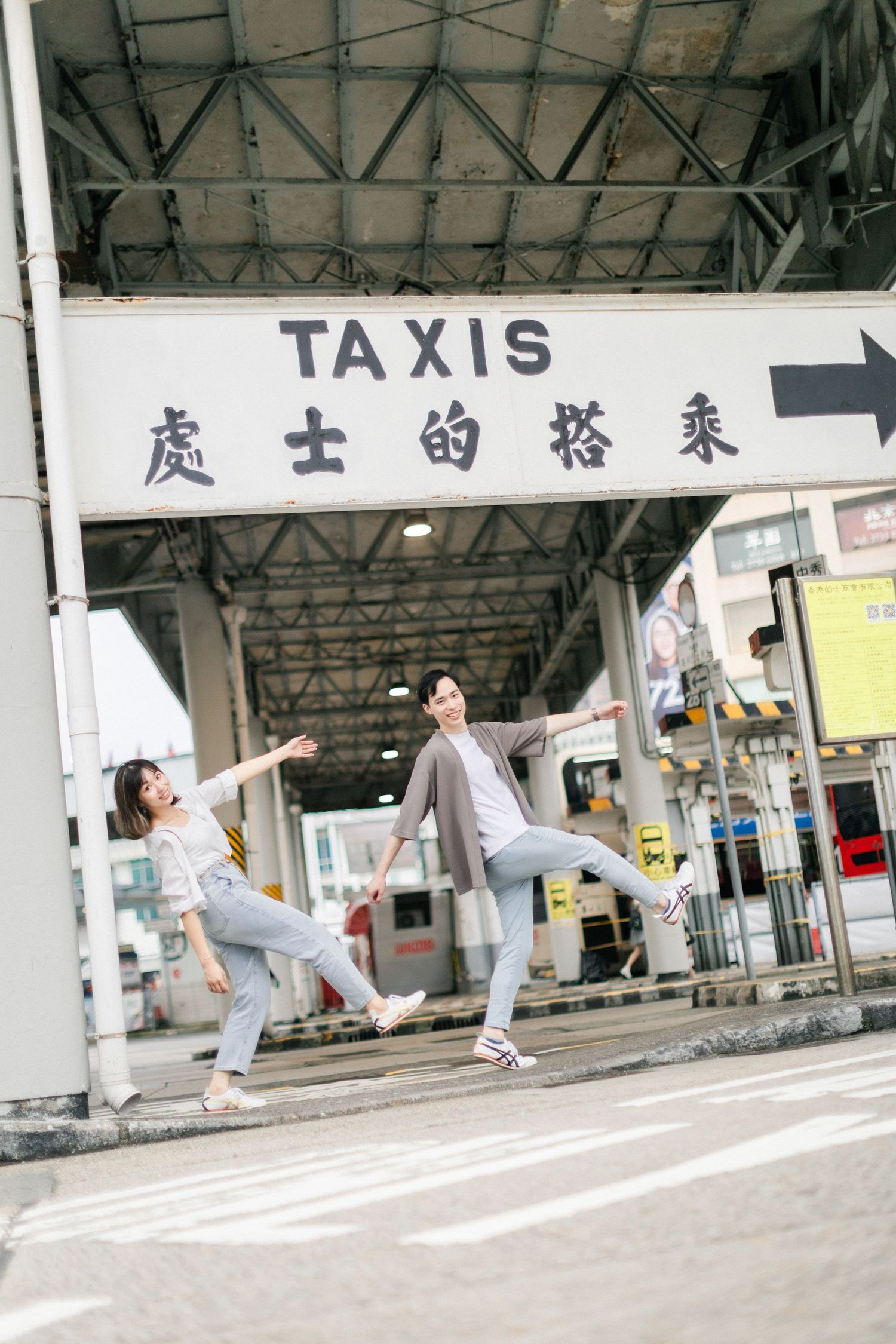 Playful Asian couple jumping and posing under a Taxis sign at a Hong Kong transportation terminal.