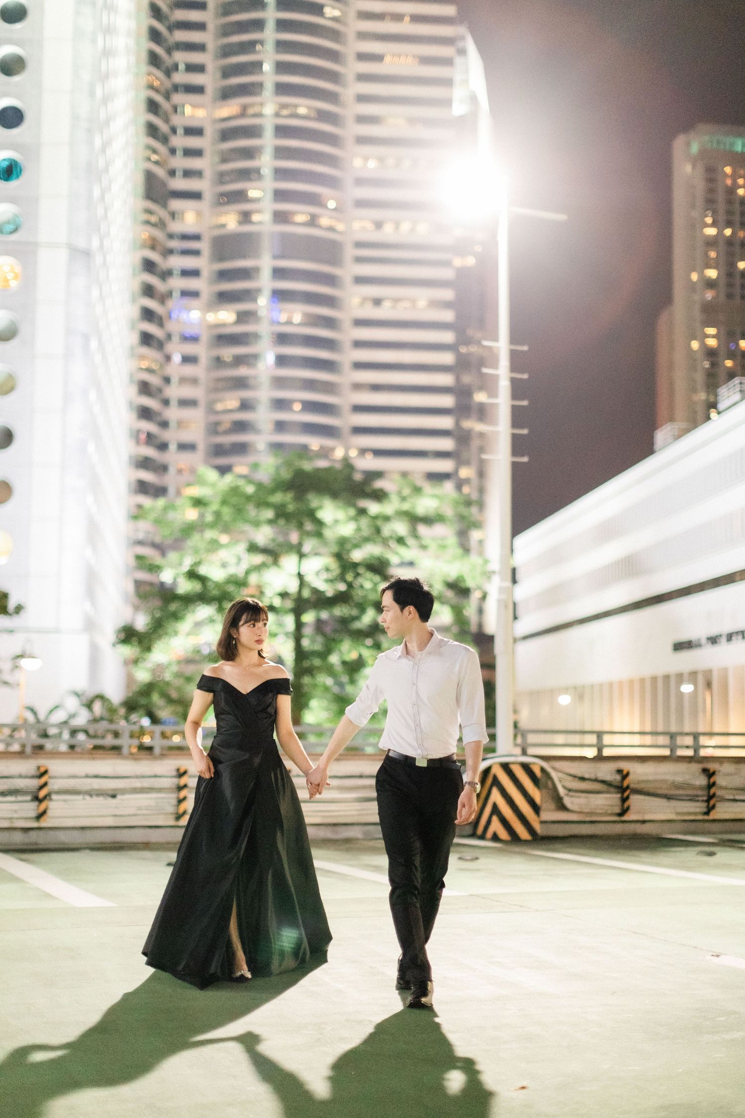 Elegant couple holding hands and walking in a city at night, woman wearing a black off-shoulder gown.