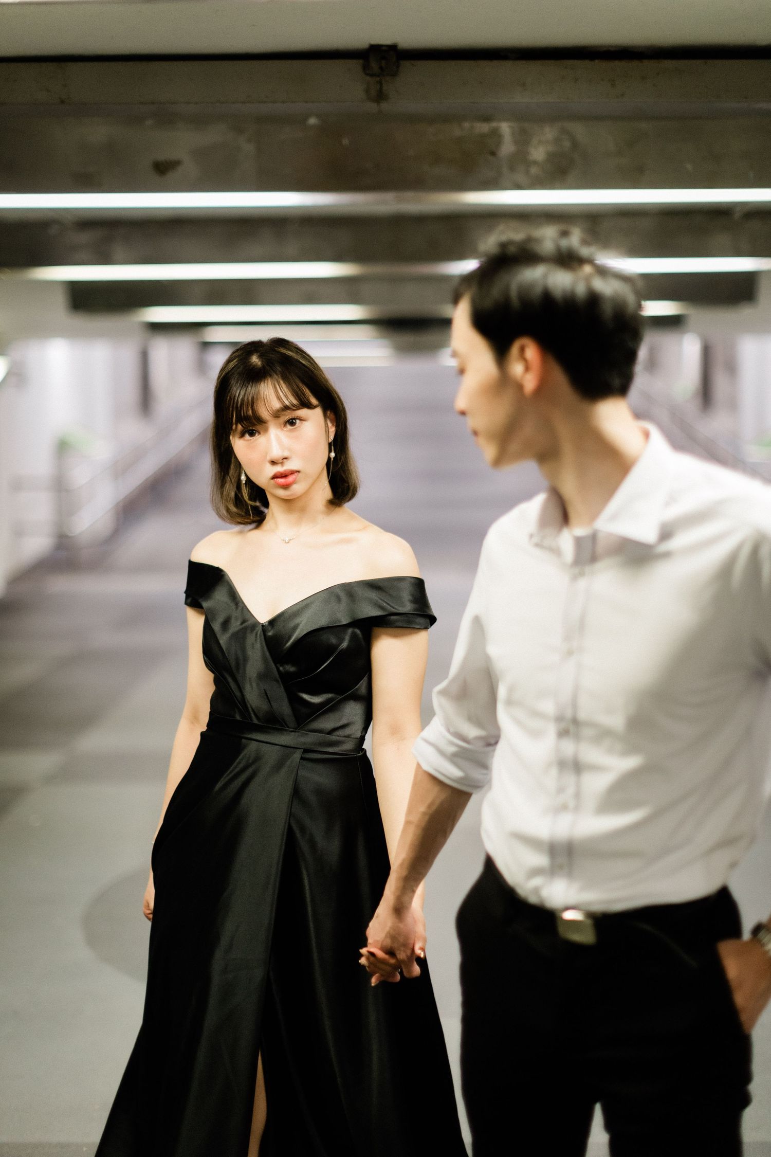 Stylish Asian woman in black off-shoulder dress holding hands with man in white shirt in a parking garage.