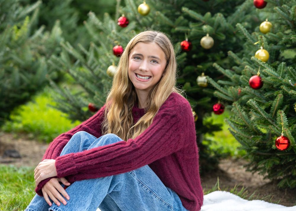A girl poses in burgundy sweater and jeans during family photo session at Mistletoe Christmas Tree Farm in Stow, MA